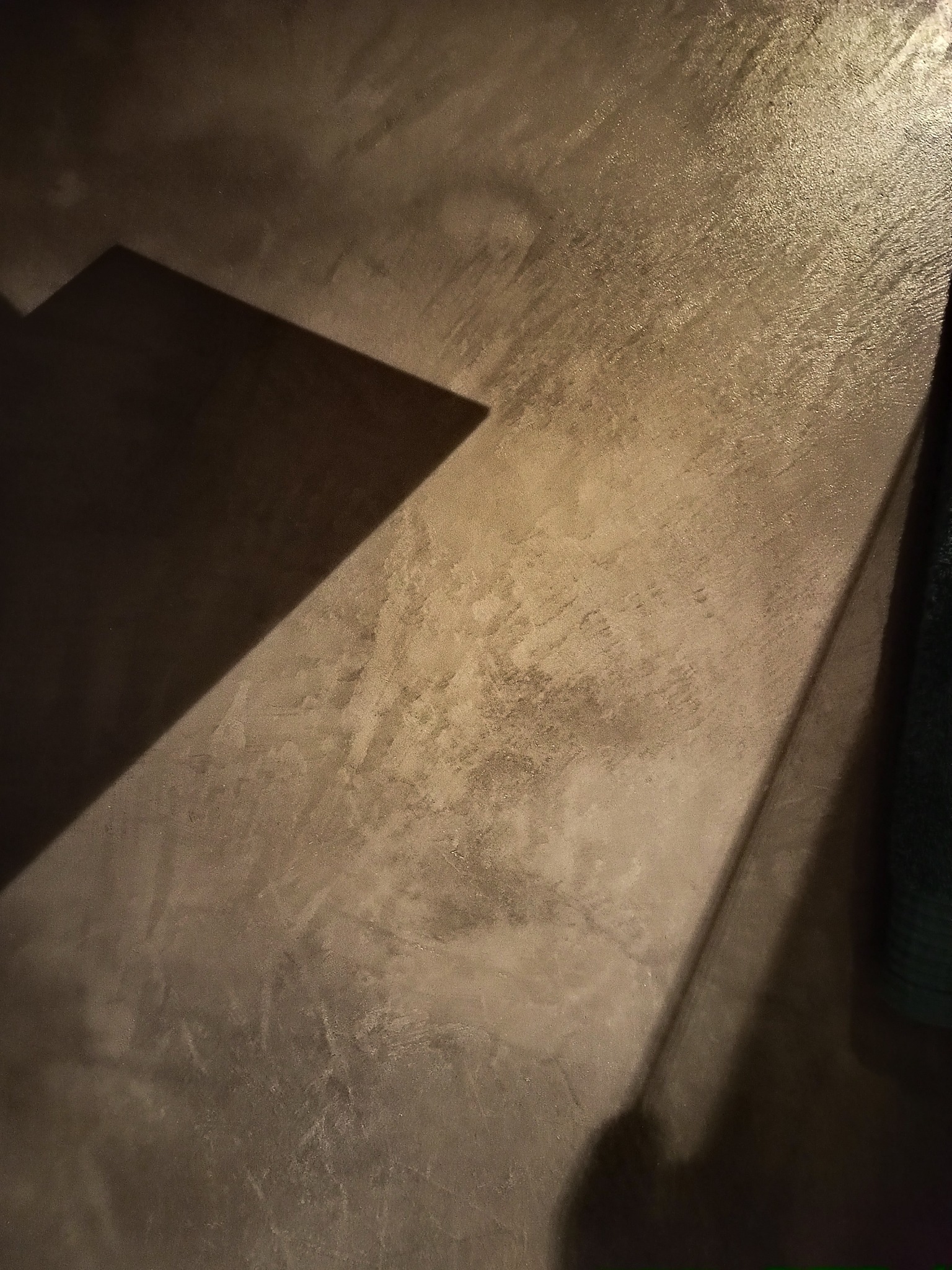 Szara, lekko błyszcząca powierzchnia podłogi z widocznym cieniem rzucanym przez niezidentyfikowany obiekt, oświetlona z góry, z fragmentem zielonego materiału w prawym dolnym rogu.