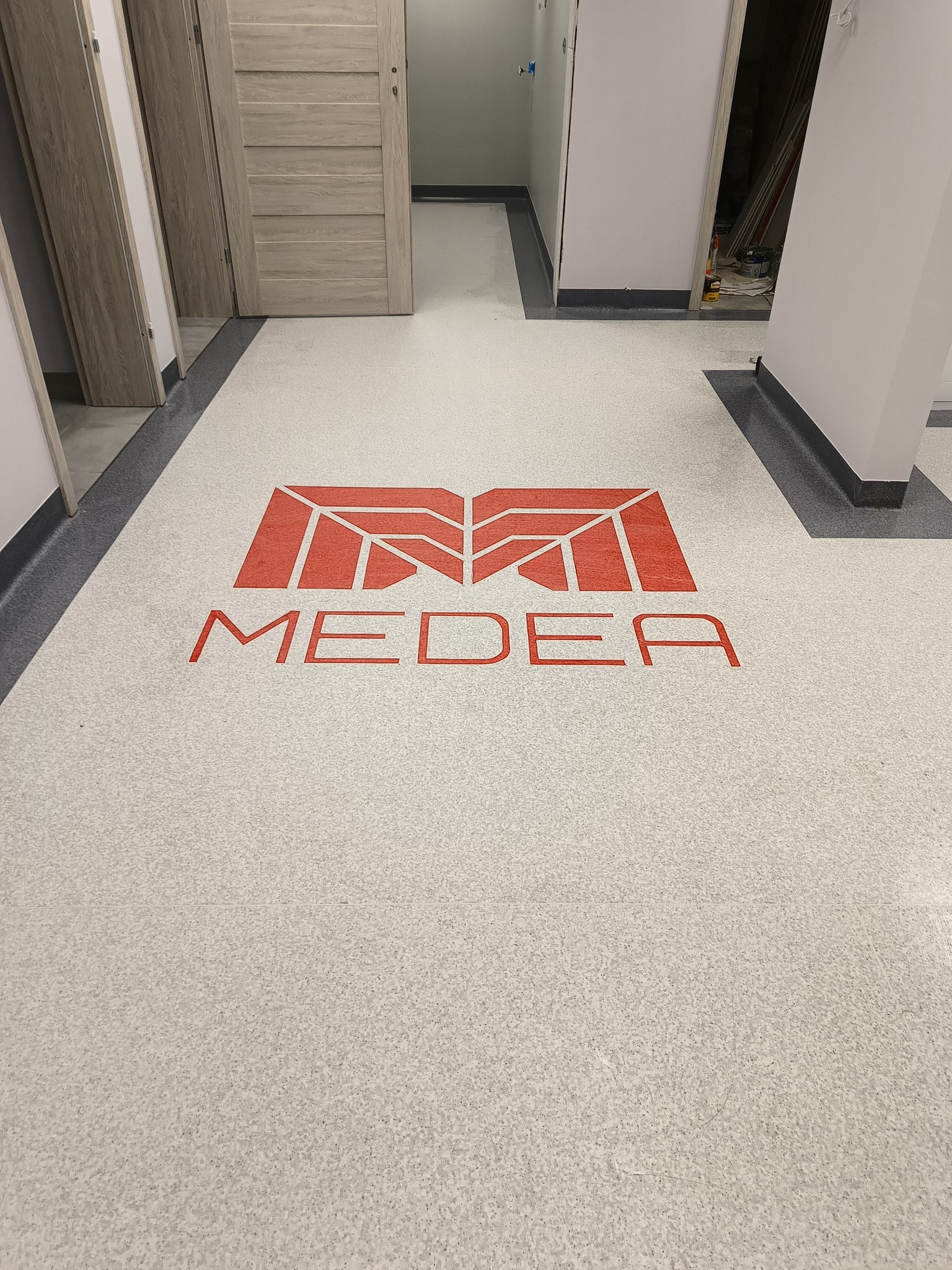 Jasnoszara podłoga w nowym budynku z czerwonym logo 'MEDEA' w centralnej części, widoczne drzwi i fragmenty korytarza.