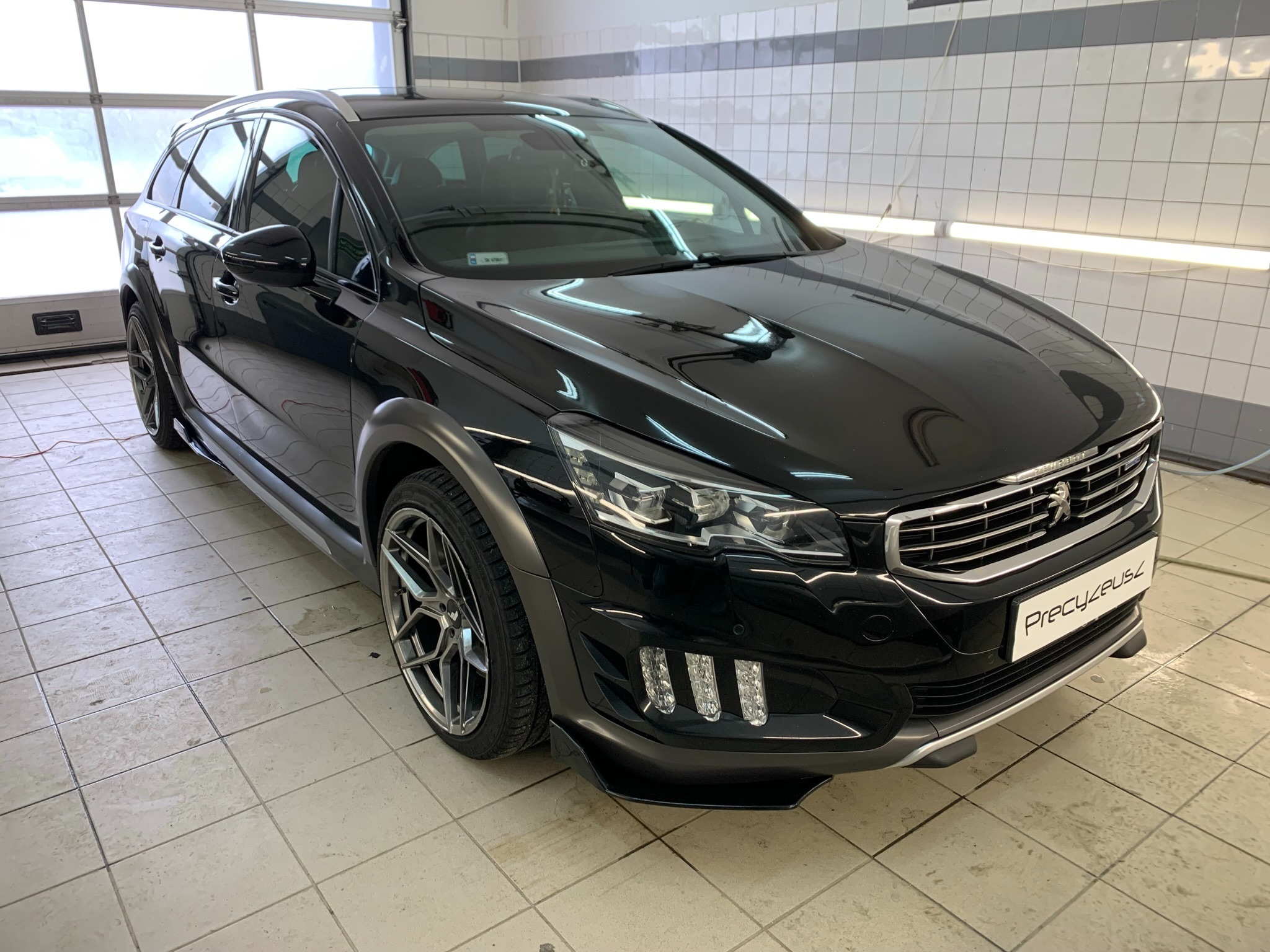 Błyszczący, czarny Peugeot kombi po detailingu, zaparkowany w czystym garażu z białymi kafelkami, widoczne logo firmy detailingowej na przednim zderzaku.
