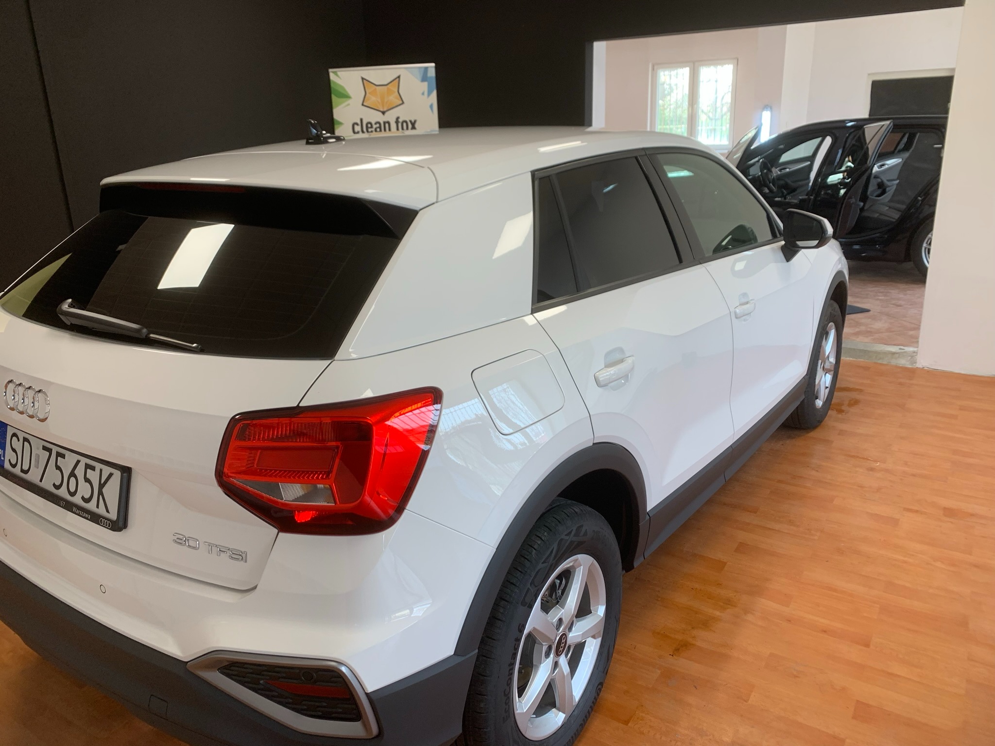 Białe Audi Q2 z przyciemnionymi szybami, widoczne tablice rejestracyjne SD 7565K oraz logo firmy Clean Fox na dachu, zaparkowane w jasnym pomieszczeniu.
