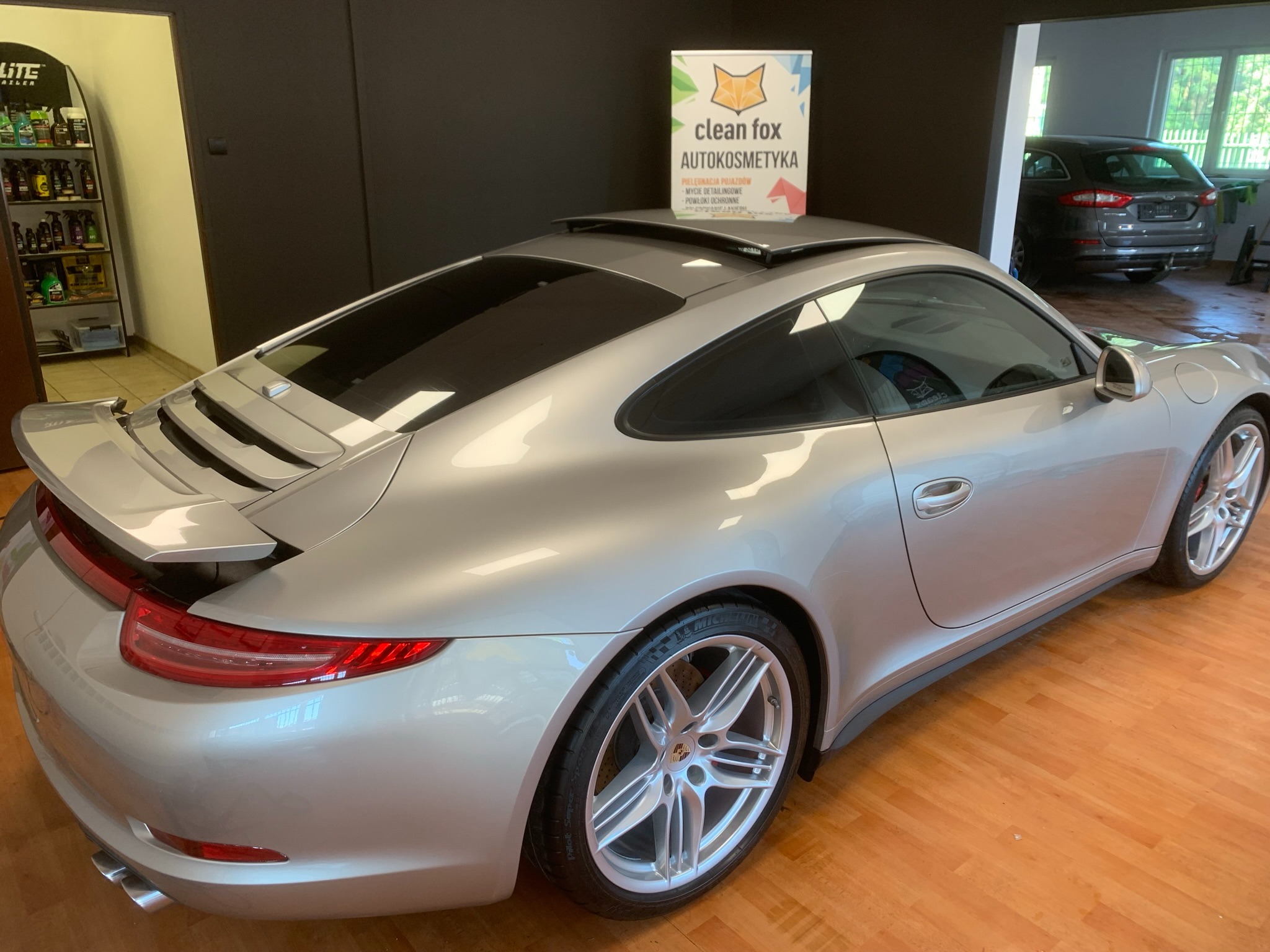 Srebrne Porsche 911 z otwartym szyberdachem, widok z tyłu w salonie detailingu, z logo Clean Fox Autokosmetyka na dachu i półkami z kosmetykami w tle.