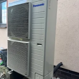 Zewnętrzna jednostka pompy ciepła Panasonic, zamontowana na betonowych blokach przed ścianą budynku z widoczną rurą odprowadzającą skropliny.