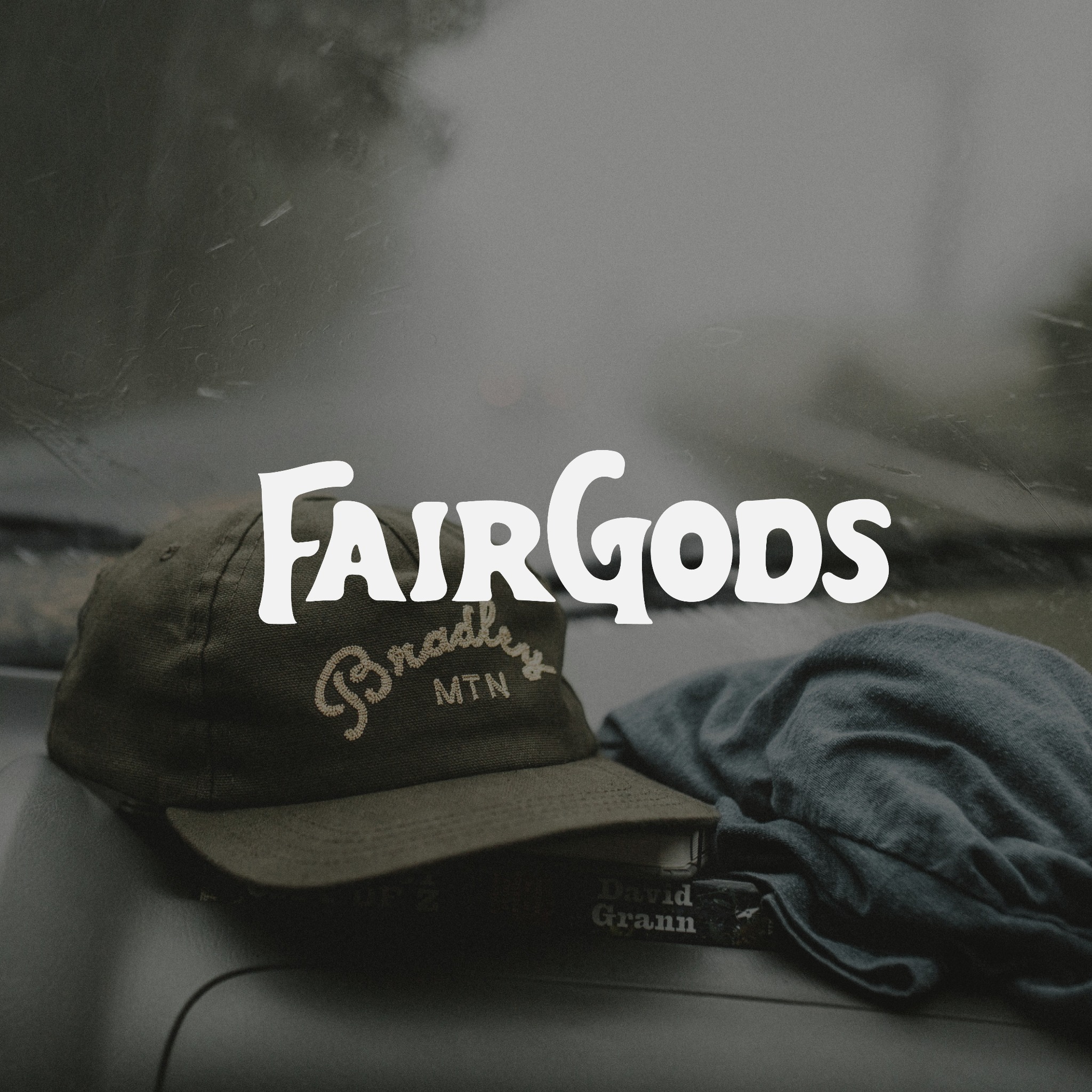 Czapka z daszkiem z napisem 'Bradley MTN' leżąca na desce rozdzielczej samochodu obok książki Davida Granna, z logo 'FairGods' nałożonym na zdjęcie i zamglonym tłem.