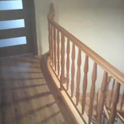 Jasne drewniane poręcze schodowe z okrągłymi szczeblami, rzucające cienie na drewnianą podłogę, obok drzwi z przeszkleniami.