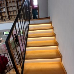 Henryk Woźniak - Nowoczesne schody z podświetleniem LED i czarną metalową balustradą, widok z dołu, wnętrze domu z antresolą w tle.
