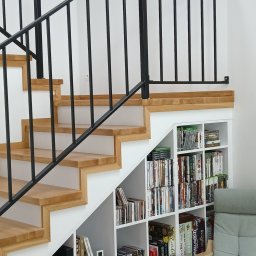 Henryk Woźniak - Nowoczesne schody z drewnianymi stopniami i czarną metalową balustradą. Pod schodami białe regały z książkami i grami. Jasne wnętrze.