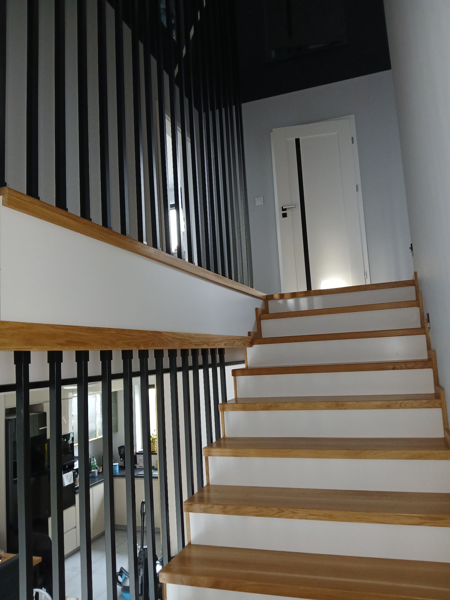 Nowoczesne schody z drewnianymi stopniami i czarną, pionową balustradą, widok z dołu. W tle białe drzwi i fragment kuchni.