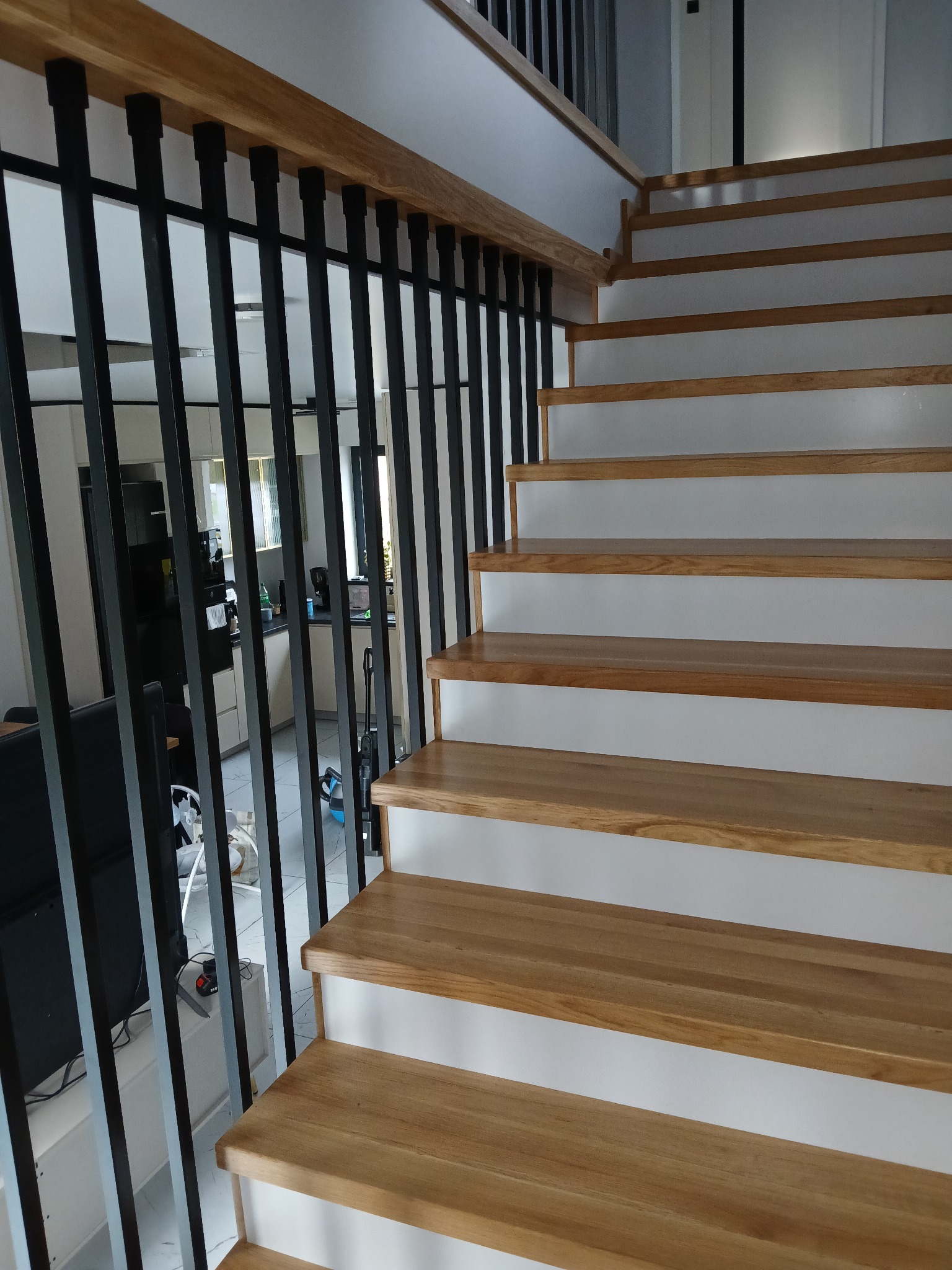 Nowoczesne schody z drewnianymi stopniami i czarną, metalową balustradą. Widok z dołu, na tle jasnego wnętrza domu z widoczną kuchnią.