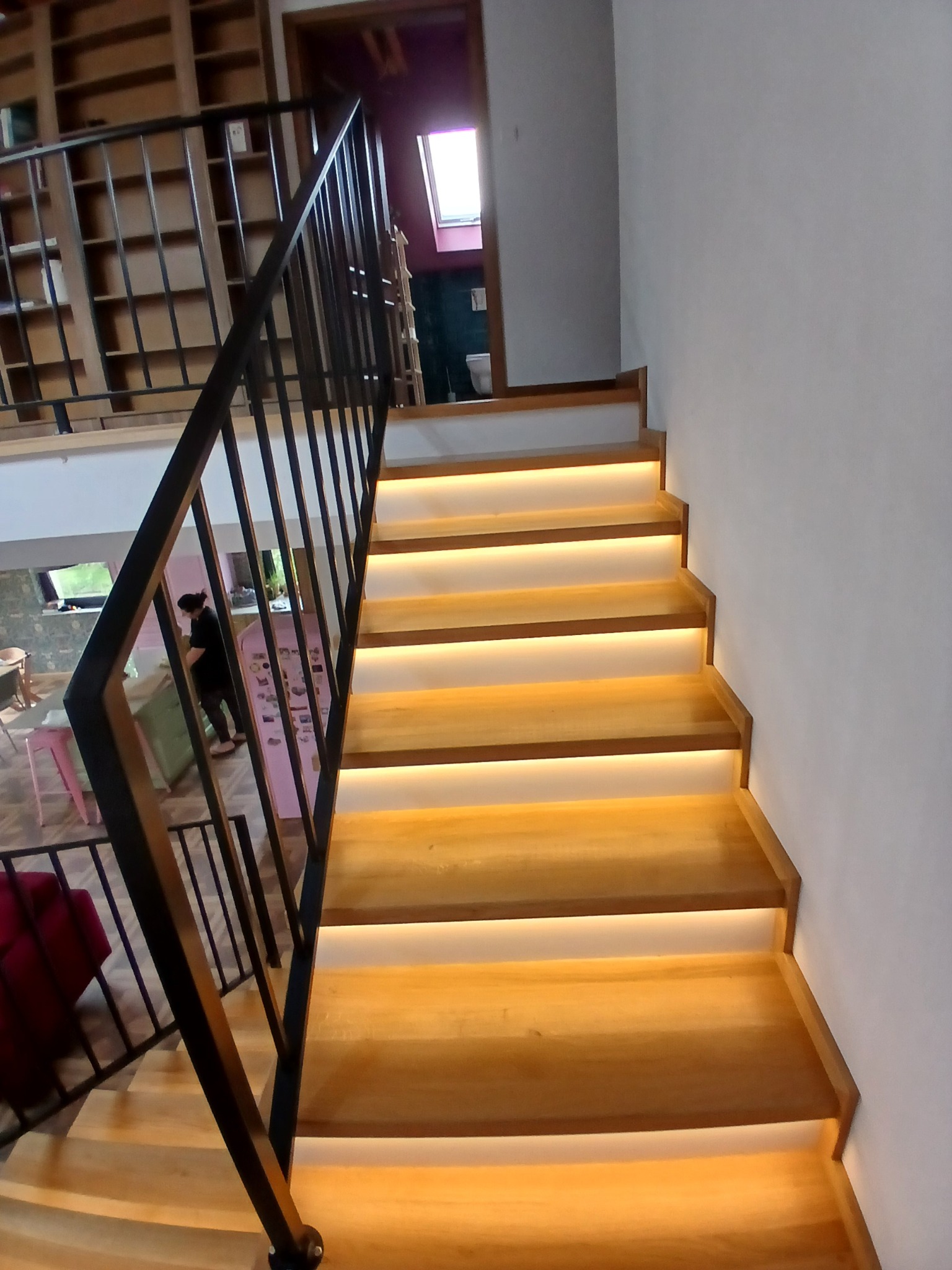 Nowoczesne schody z podświetleniem LED i czarną metalową balustradą, widok z dołu, wnętrze domu z antresolą w tle.