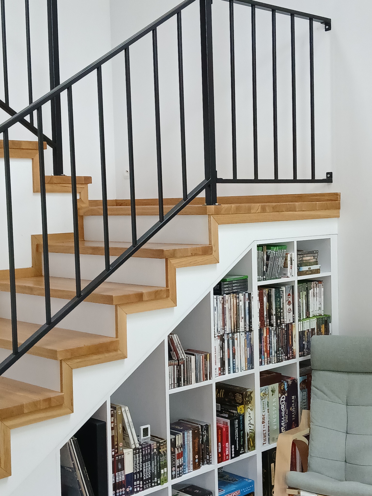 Nowoczesne schody z drewnianymi stopniami i czarną metalową balustradą. Pod schodami białe regały z książkami i grami. Jasne wnętrze.