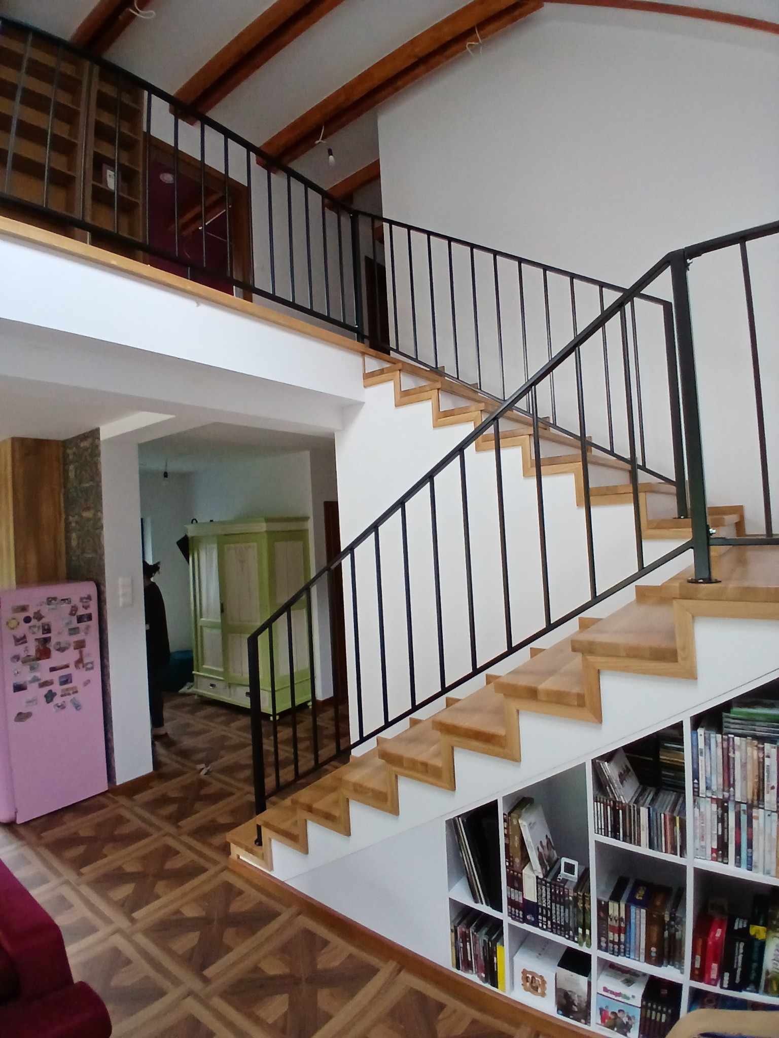 Nowoczesne schody z jasnego drewna z czarną metalową balustradą, wkomponowane w przestrzeń z regałami na książki pod spodem. Widok z dołu na antresolę.