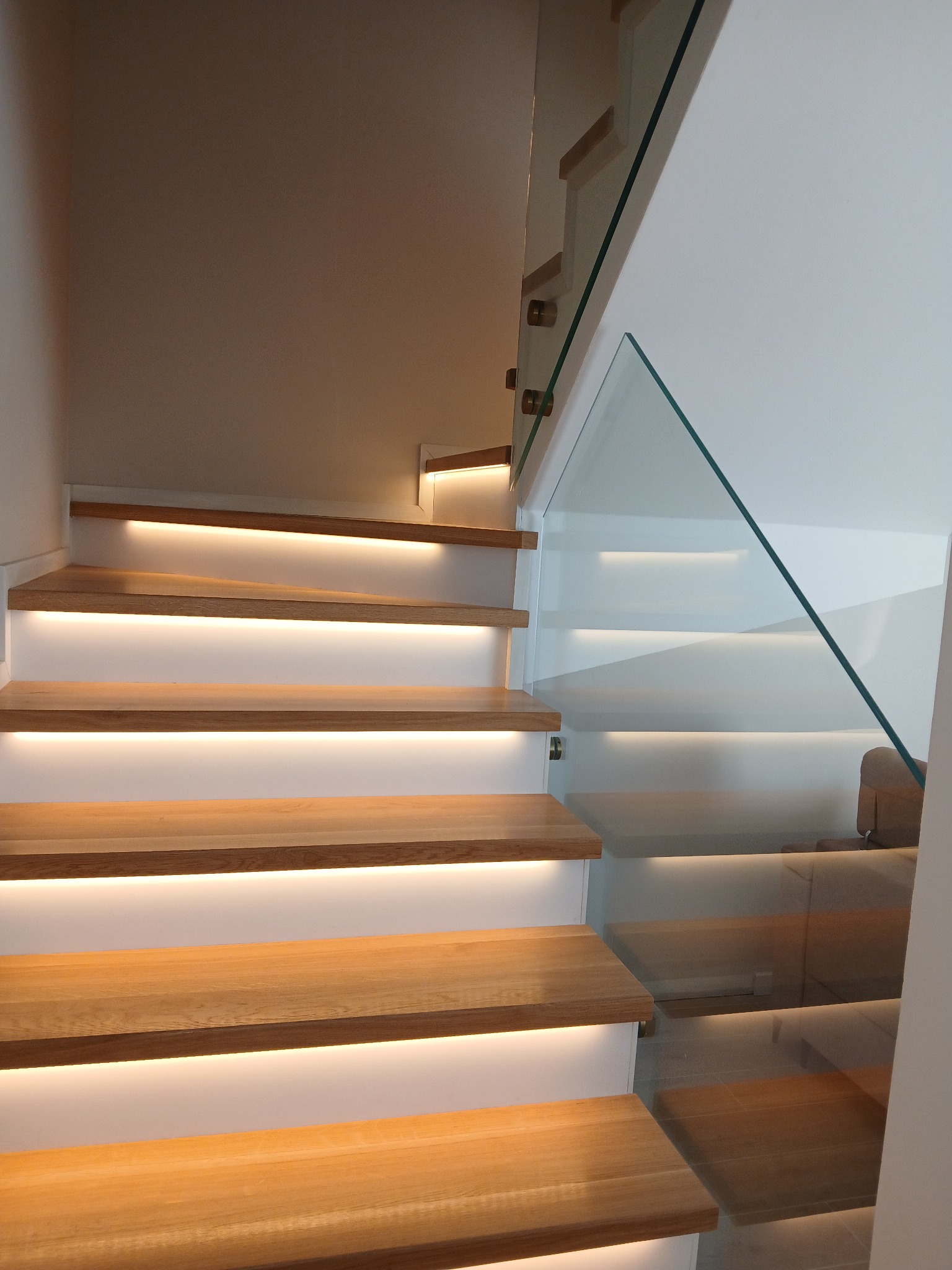 Nowoczesne schody drewniane z podświetleniem LED i szklaną balustradą. Jasne drewno, białe stopnie, minimalistyczny design. Widok z dołu, oświetlenie podkreśla detale.