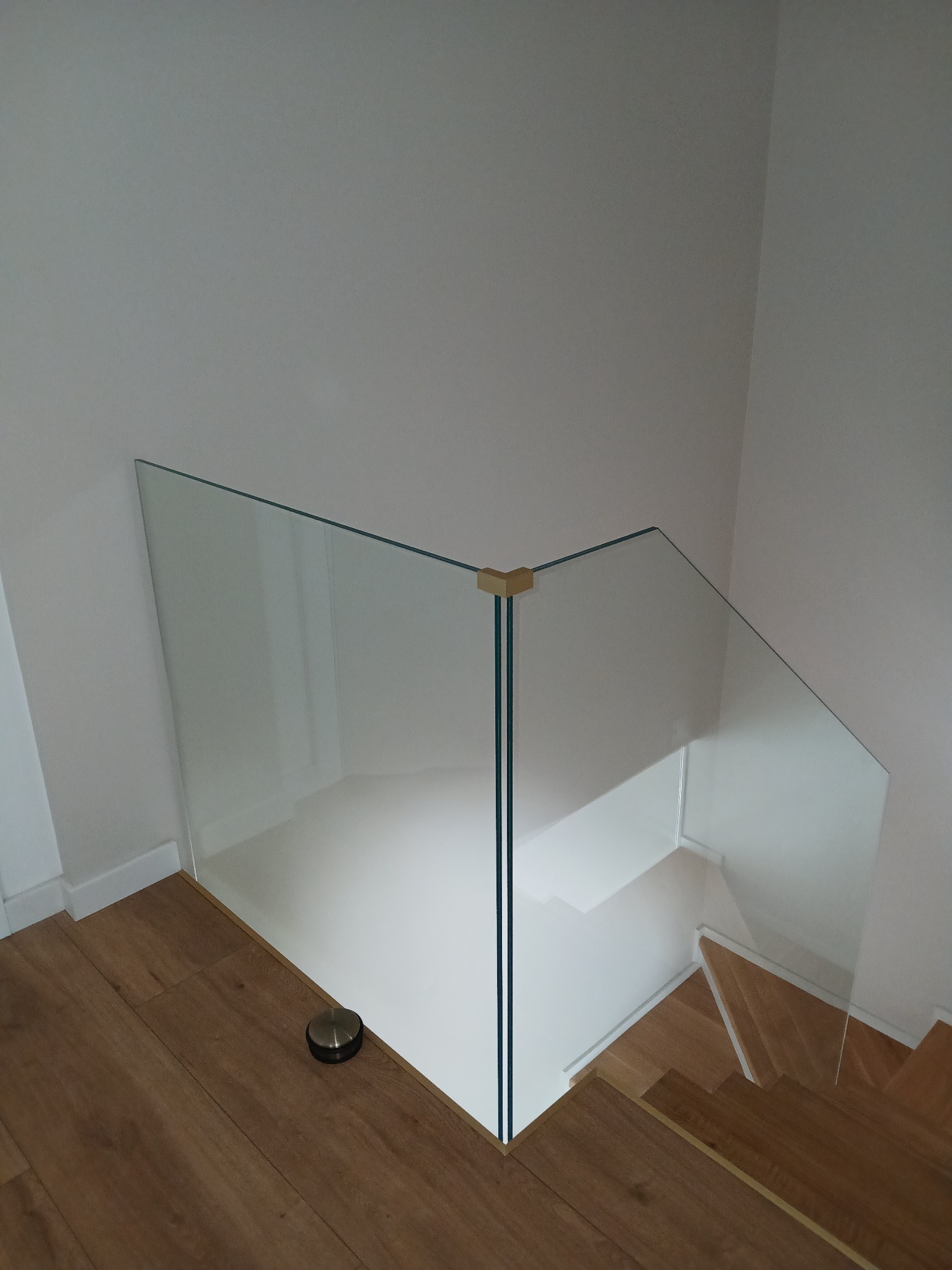 Szklana balustrada zabezpieczająca schody z drewnianymi stopniami, widok z góry, minimalistyczny design z elementami mocującymi w kolorze złotym.