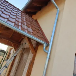 Nowa, metalowa rynna spustowa zamontowana na elewacji domu z brązową dachówką ceramiczną i drewnianą konstrukcją zadaszenia nad wejściem, drabina oparta o budynek.