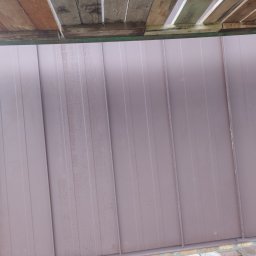 Usługi dekarskie ciesielskie i ogólnobudowlane - Nowe, brązowe panele blaszane na dachu, widoczne ze rusztowania. Świeżo położone, z delikatnymi śladami wilgoci. Ujęcie z dołu, podkreślające regularność i estetykę.