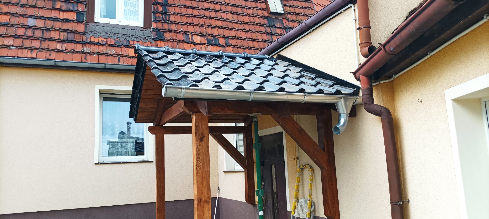 Zadaszenie nad wejściem do domu z drewnianą konstrukcją wspartą na słupach, pokryte czarną dachówką ceramiczną z zamontowaną rynną. W tle widoczny fragment dachu domu z czerwoną dachówką karpiówką.