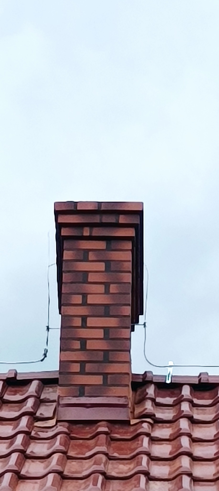 Ceglany komin na dachu z dachówki ceramicznej w pochmurny dzień, widoczne przewody antenowe.