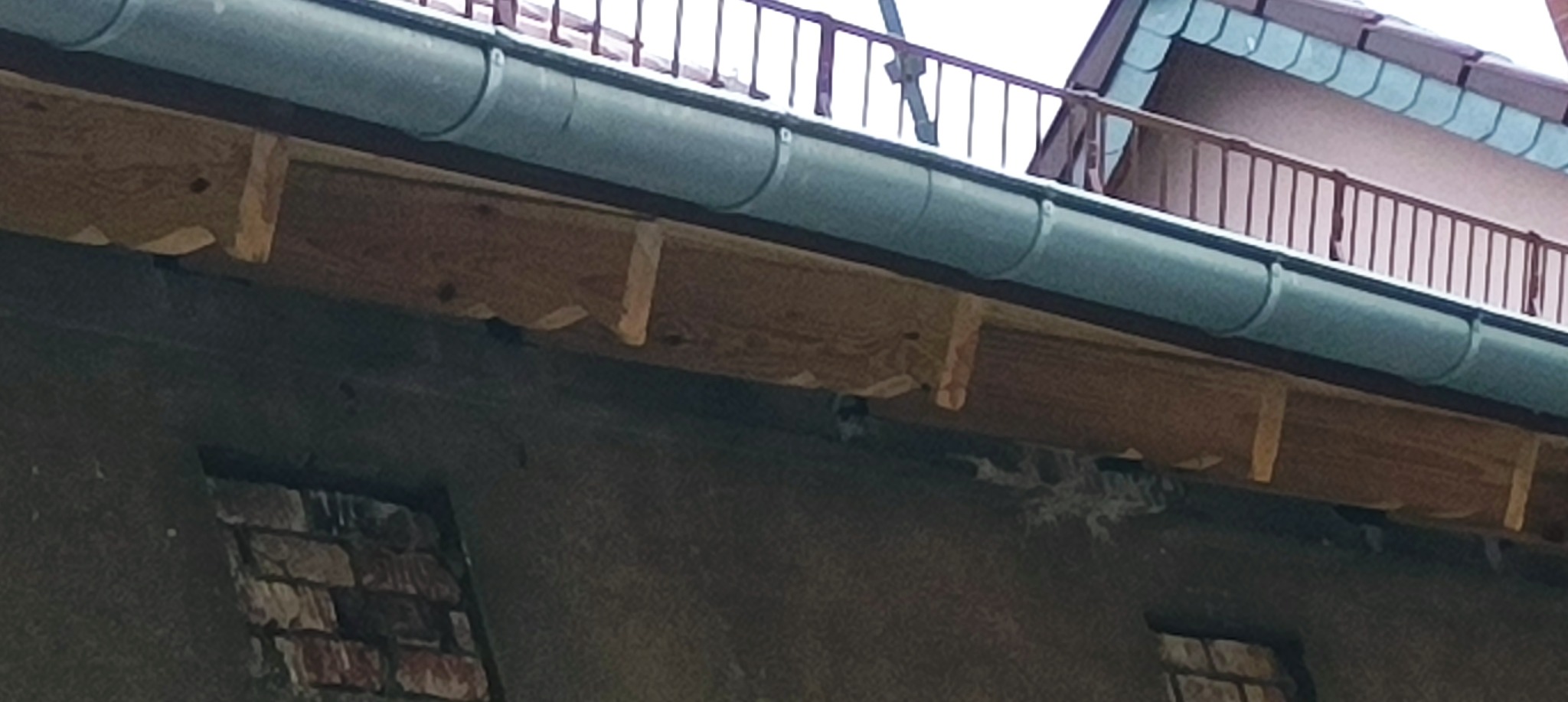 Widok z dołu na zamurowane okna, drewnianą konstrukcję podtrzymującą zieloną rynnę i fragment dachu z metalowymi płytkami w tle.