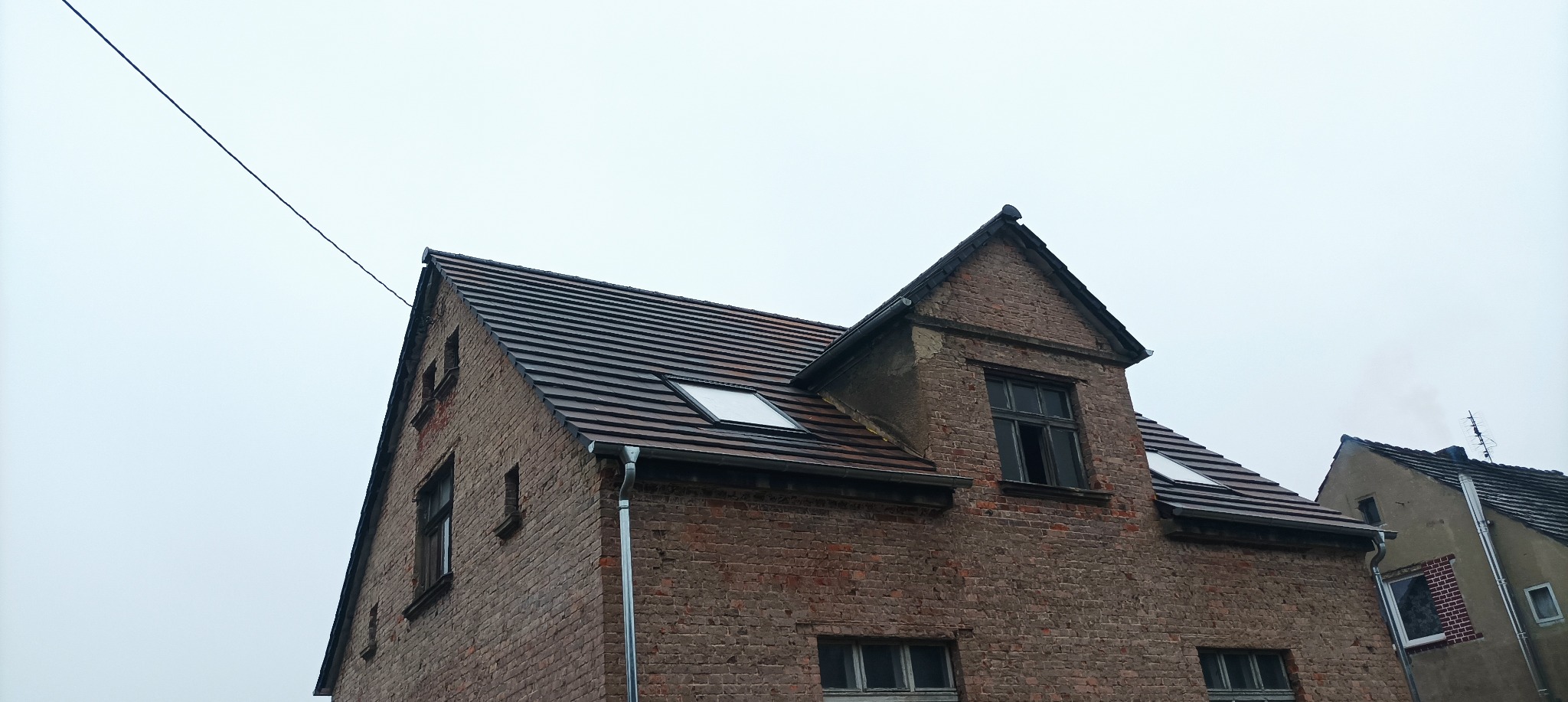 Ceglany budynek z ciemną dachówką w Lubsku, widoczne okna dachowe i rynna, pochmurne niebo w tle.