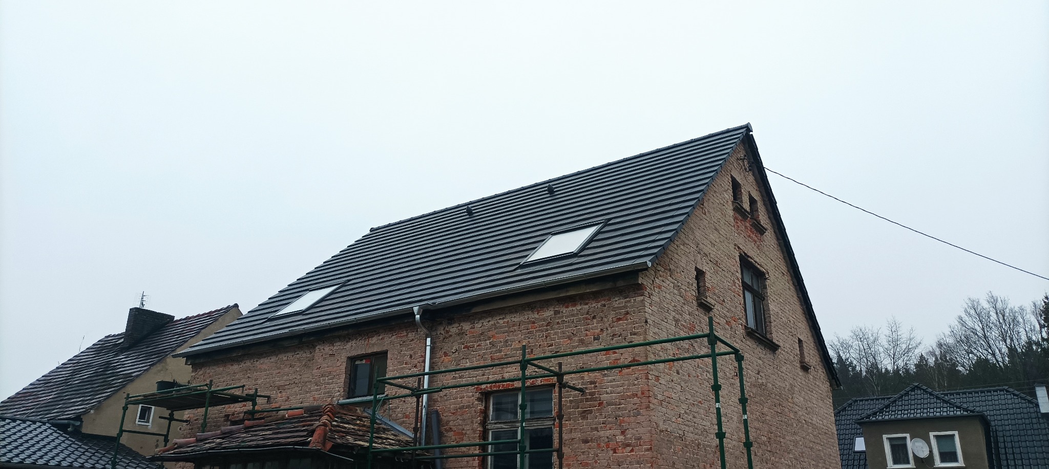 Dom z czerwonej cegły z nowym, ciemnoszarym dachem z dachówką ceramiczną i dwoma oknami dachowymi, widoczne rusztowania.