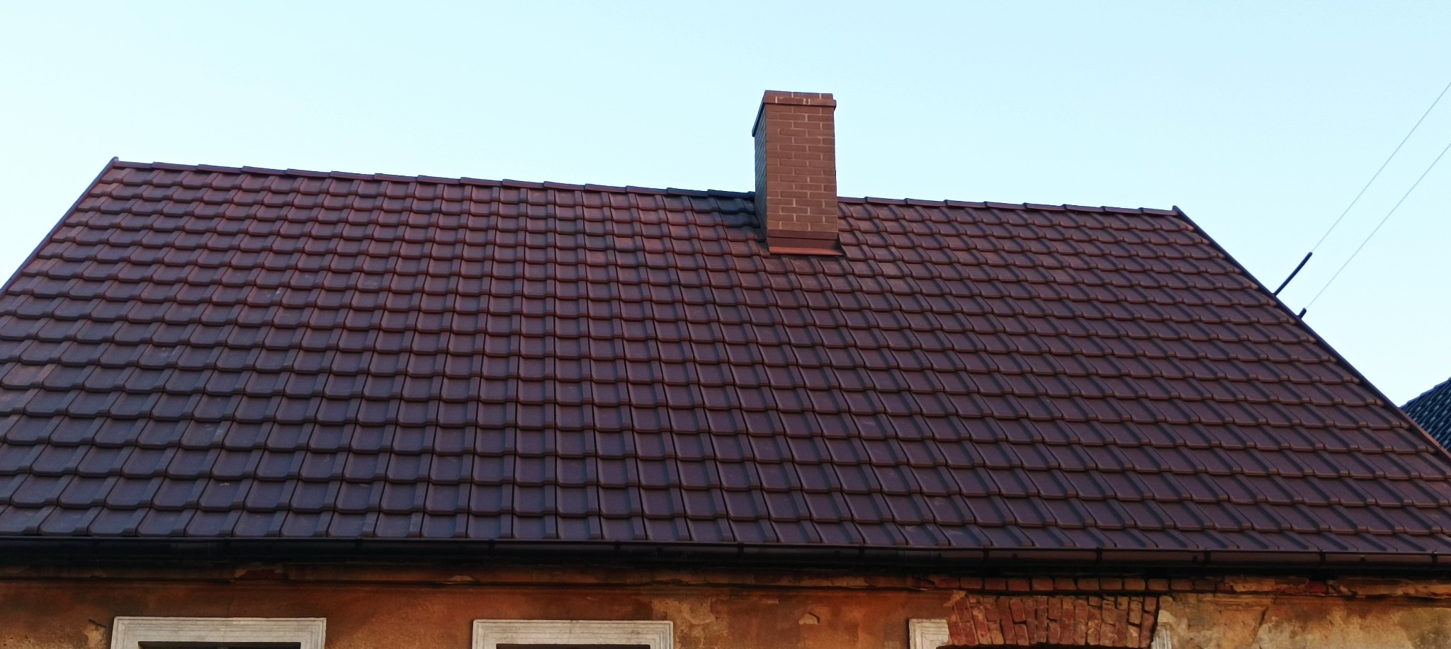 Ciemnobrązowa dachówka ceramiczna na dachu domu z ceglanym kominem, widoczne okna w starej elewacji.