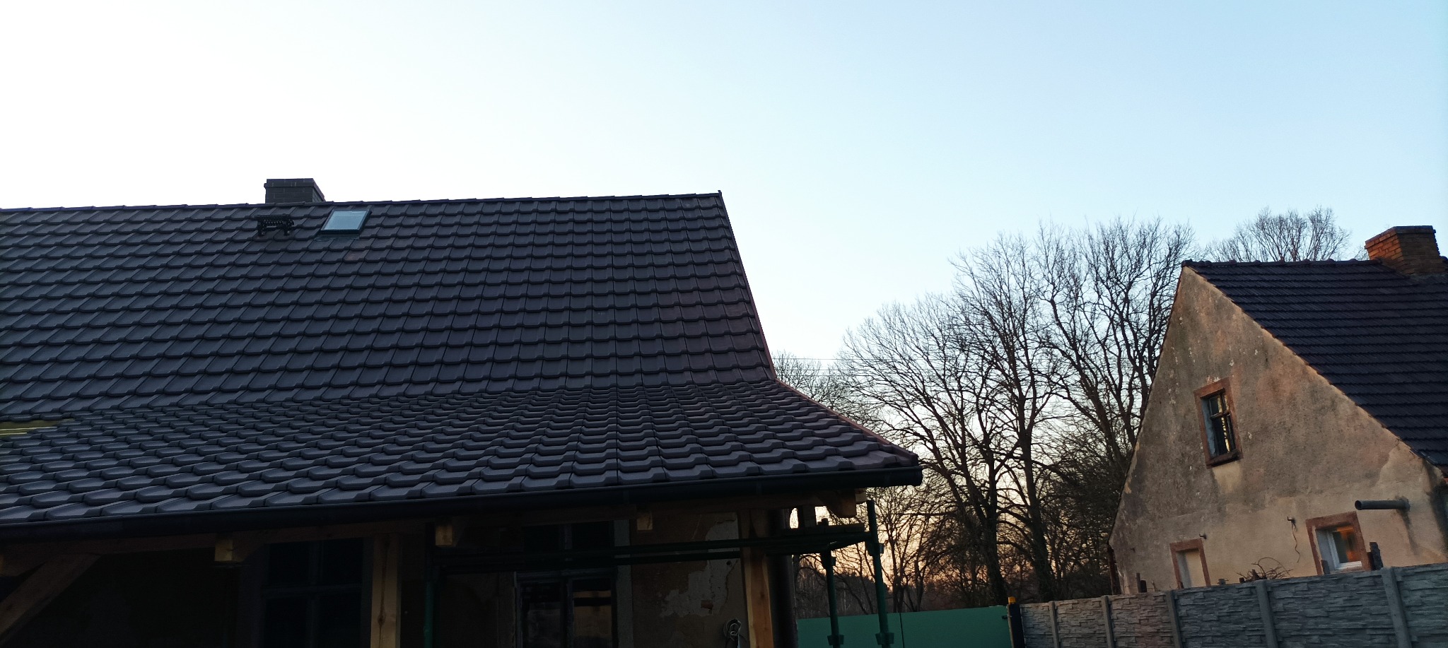 Nowy, ciemnobrązowy dach z dachówki ceramicznej z oknem dachowym i kominem, widok z dołu na tle błękitnego nieba i sąsiedniego budynku z podobnym dachem.