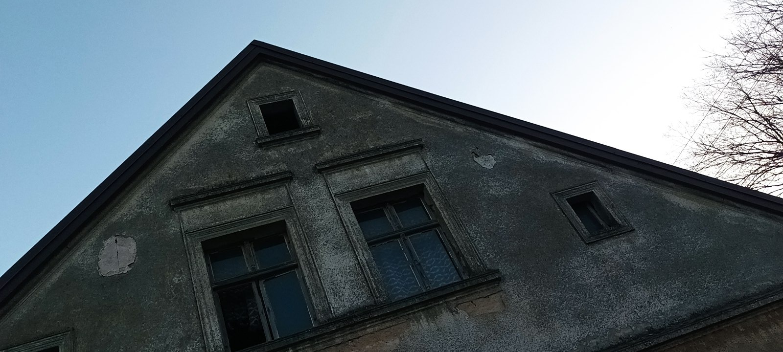 Szczyt starego, zaniedbanego domu z widocznymi uszkodzeniami tynku, małymi oknami i brakiem jednego okna na samej górze.