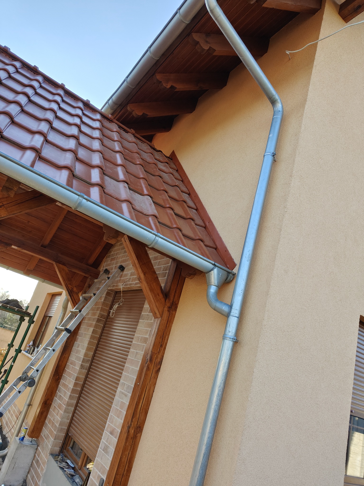 Nowa, metalowa rynna spustowa zamontowana na elewacji domu z brązową dachówką ceramiczną i drewnianą konstrukcją zadaszenia nad wejściem, drabina oparta o budynek.