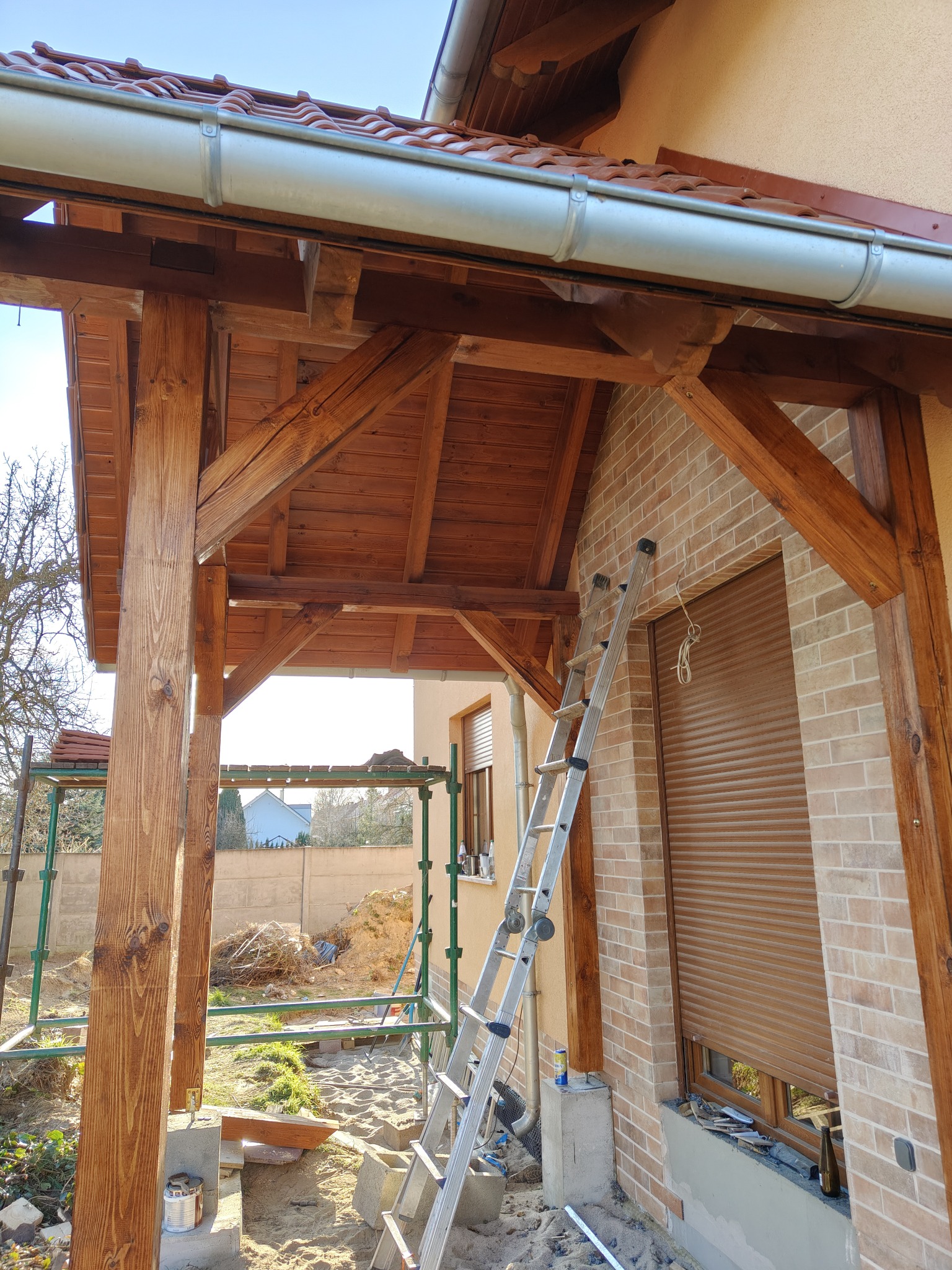 Drewniana konstrukcja z nową rynną, widoczna drabina oparta o ceglany budynek z roletą, w tle rusztowanie i ziemia.