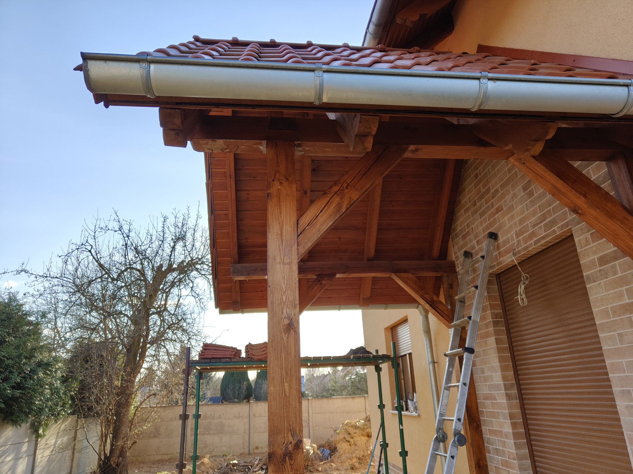 Drewniana konstrukcja z nowym dachem z dachówki ceramicznej i rynnami, widok z dołu, w tle drabina oparta o ścianę budynku z brązowej cegły.