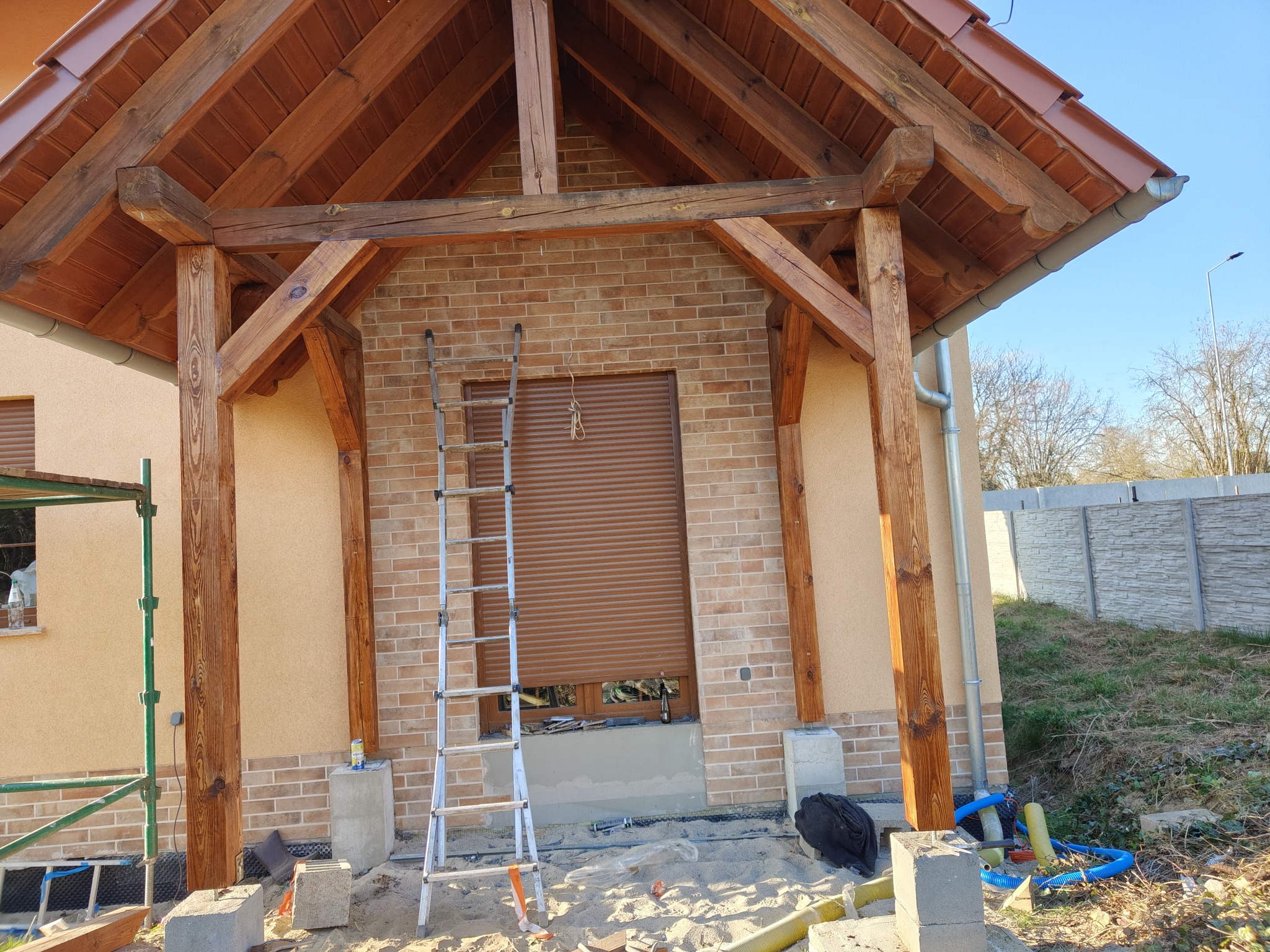 Drewniana konstrukcja z belek i słupów, prawdopodobnie weranda, w trakcie budowy, z drabina opartą o ceglaną ścianę z oknem z roletami, widoczny bałagan budowlany na piasku.