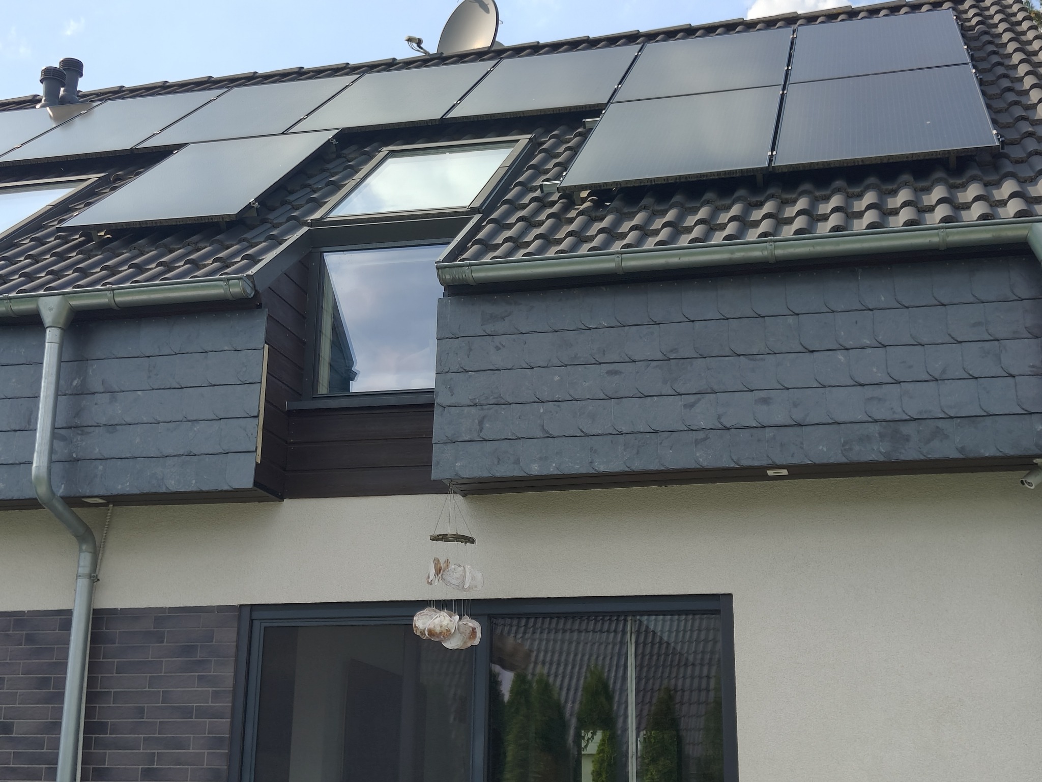 Ciemny dach z panelami słonecznymi i oknem dachowym, wykończony szarym łupkiem, z widoczną rynną i dekoracją okienną w postaci muszelek.