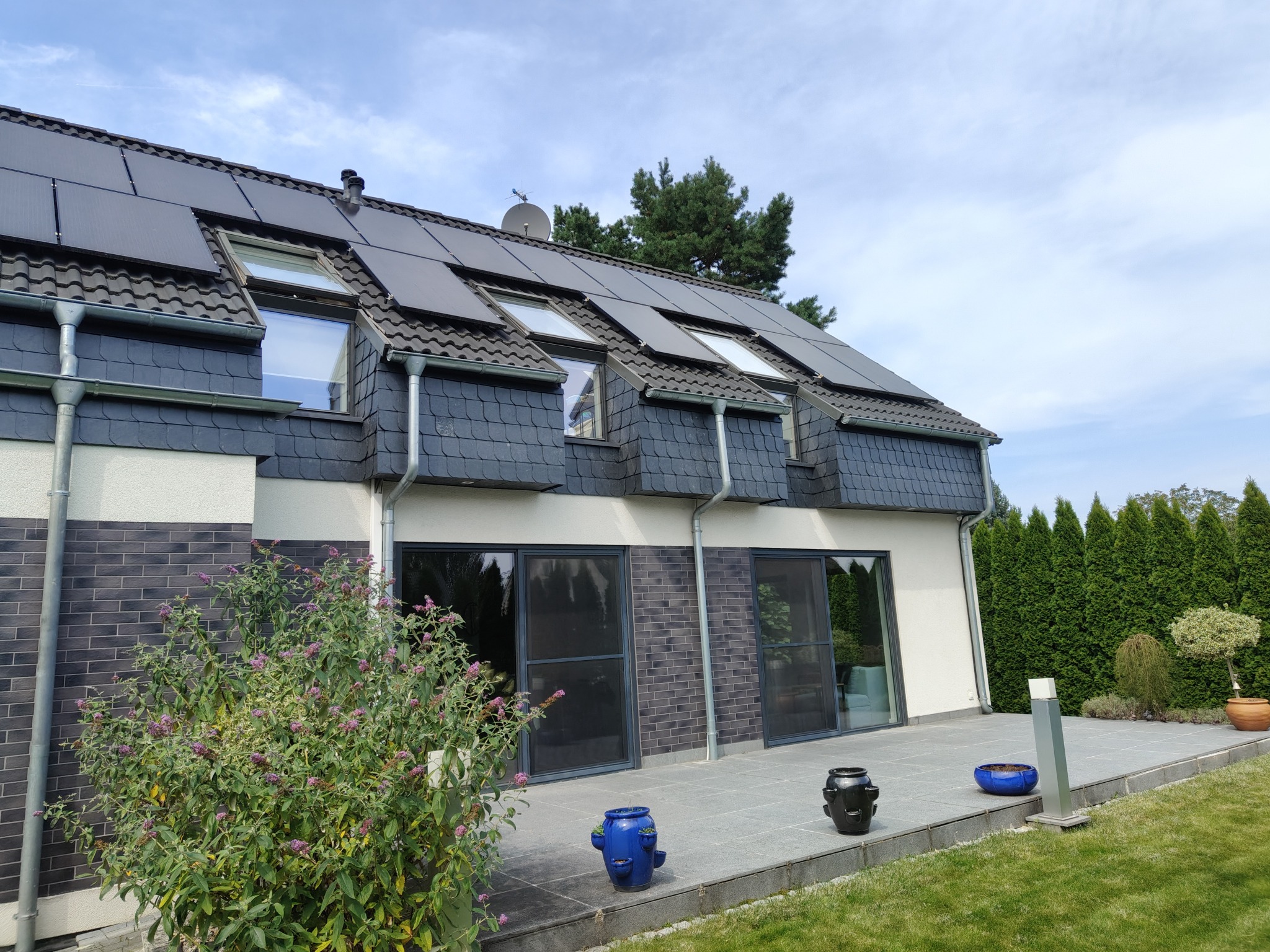 Dom z dachem pokrytym panelami słonecznymi i dachówką bitumiczną. Widoczne okna dachowe i rynny. Elewacja z elementami cegły i jasnego tynku.