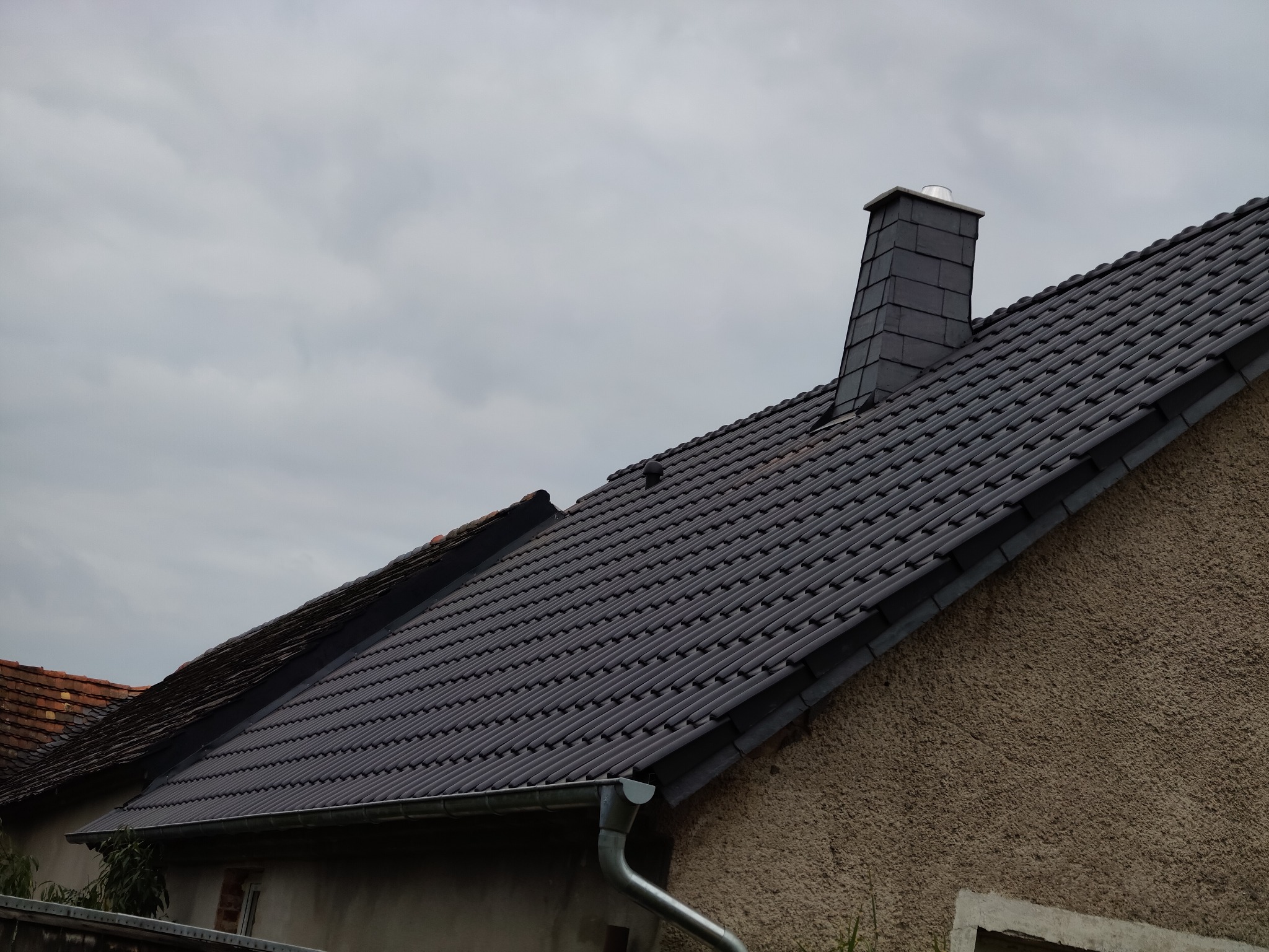 Ciemna dachówka ceramiczna na skośnym dachu domu jednorodzinnego, z widocznym kominem obłożonym łupkiem i fragmentem rynny. Pochmurne niebo w tle.