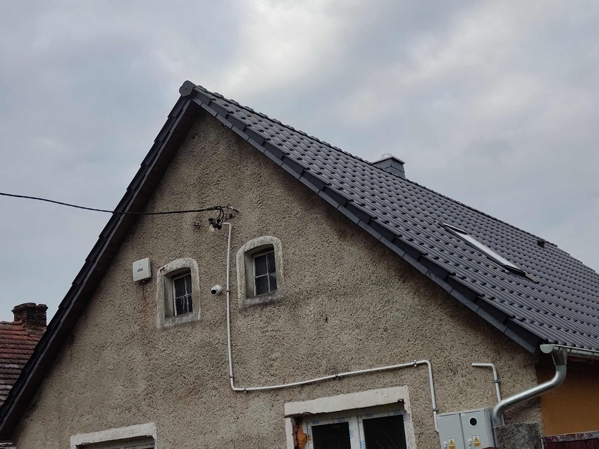 Dom z nowym, ciemnoszarym dachem z dachówki ceramicznej, widoczne okna dachowe i rynna. Elewacja w odcieniach beżu, widoczne okna i instalacje na ścianie.