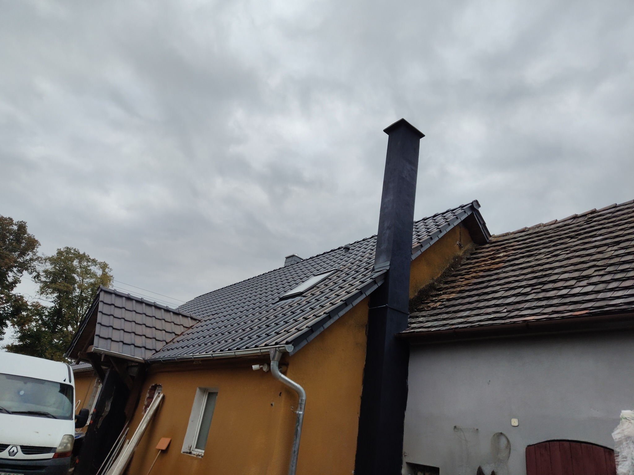 Dom z nowym, grafitowym dachem z dachówki ceramicznej i pomalowanym kominem. Widoczny fragment elewacji, rynna i okno dachowe. Pochmurne niebo w tle.
