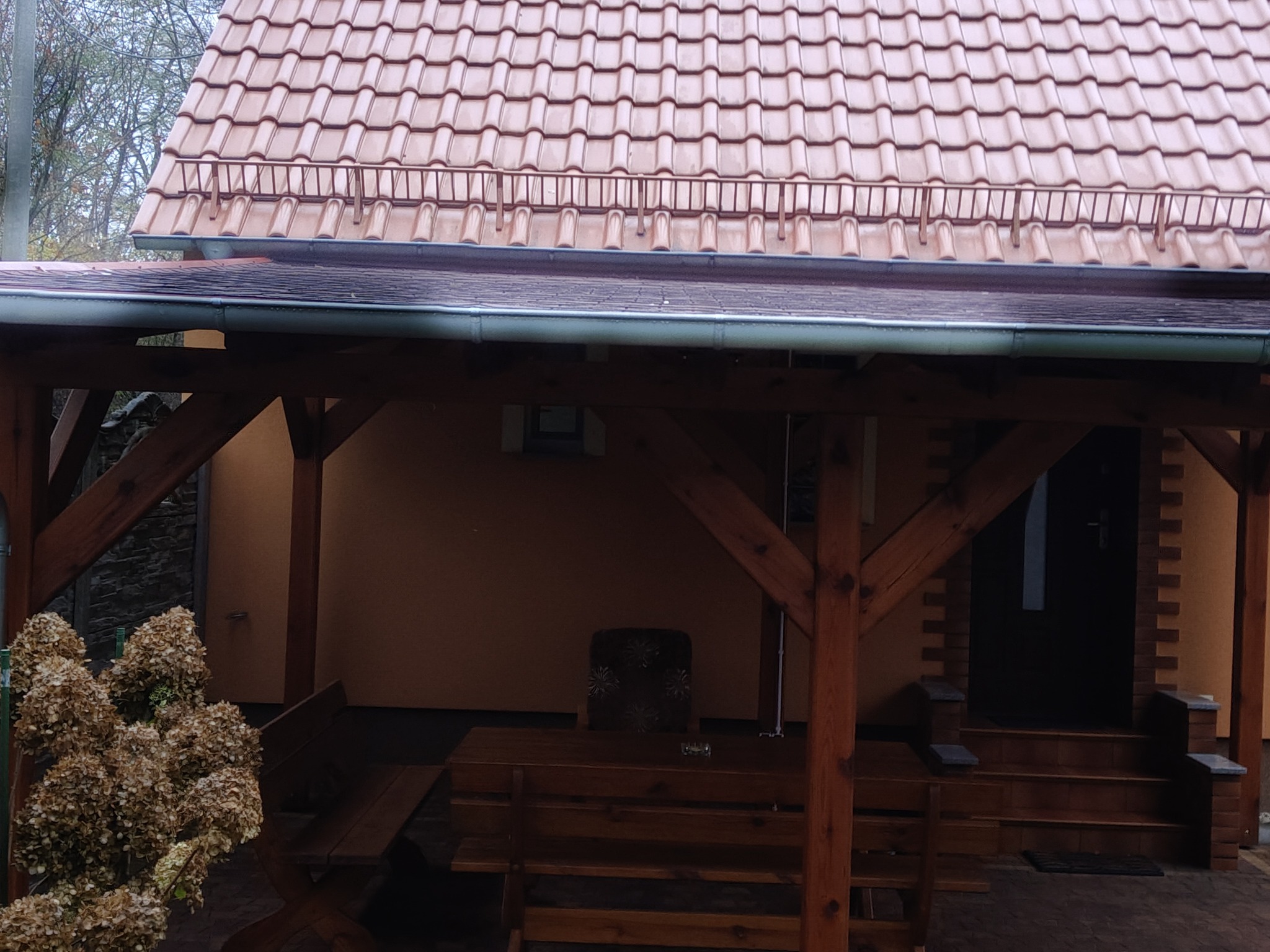 Zadaszenie z drewnianą konstrukcją i brązową dachówką. Widoczne rynny i elementy wykończenia dachu. Ujęcie z poziomu ogrodu, w tle ściana budynku.