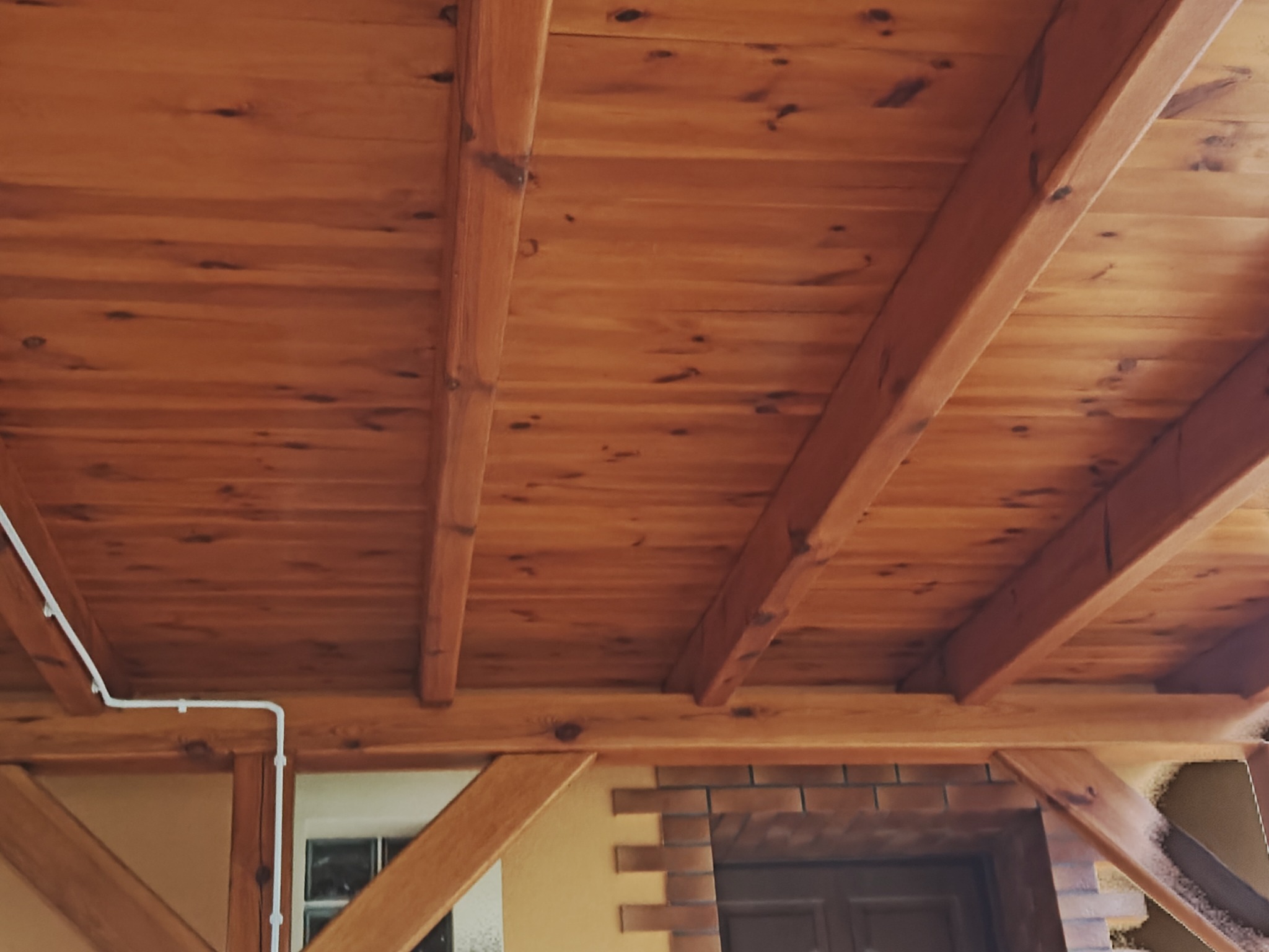 Drewniany sufit z widocznymi belkami i instalacją elektryczną, ciepłe oświetlenie, fragment ściany z ceglaną dekoracją w tle. Detal wykończenia wnętrza.