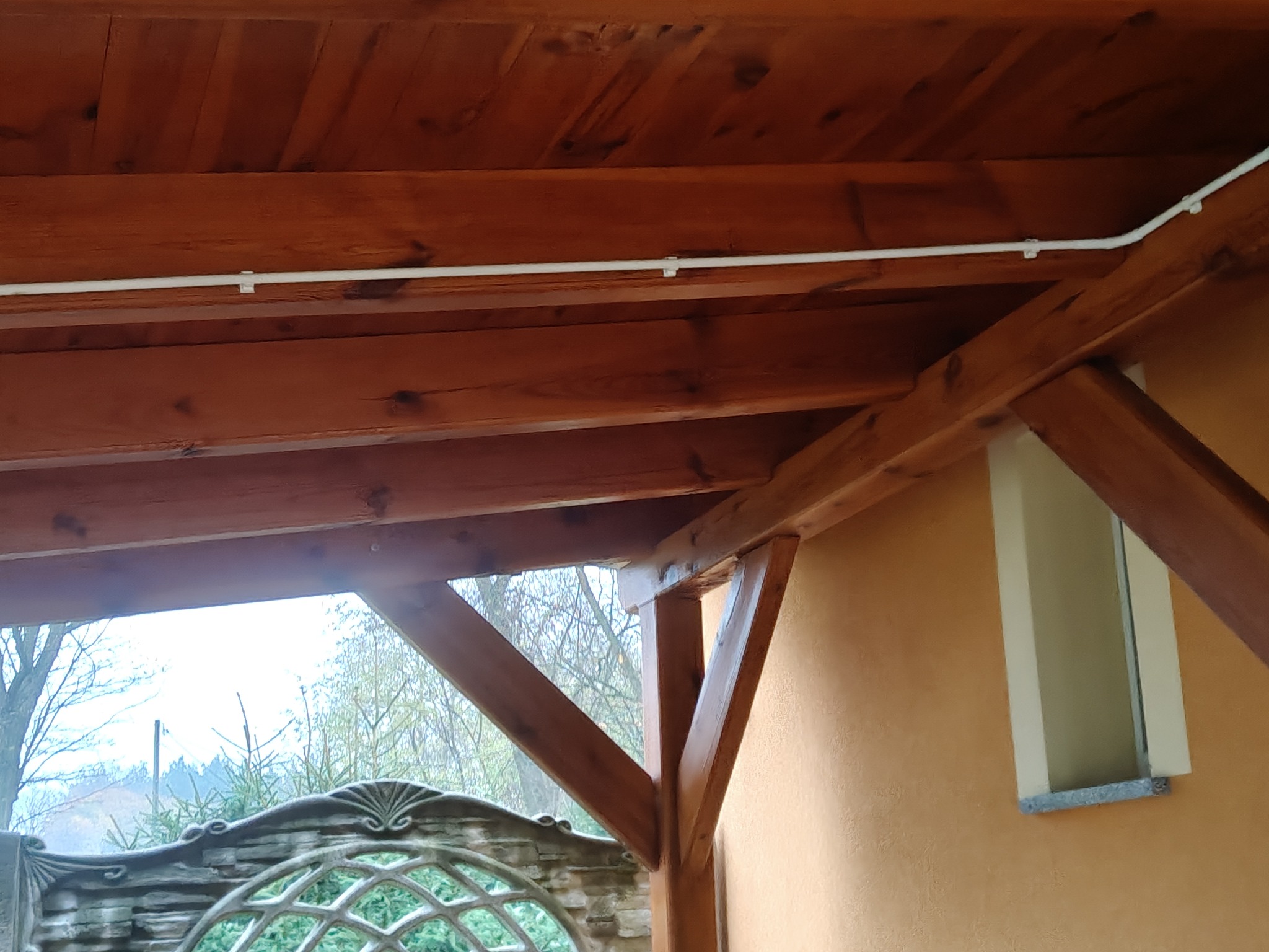 Drewniana konstrukcja dachu z widoczną instalacją elektryczną, brązowe belki, widok na zewnątrz przez ozdobne ogrodzenie. Ujęcie z dołu, ciepłe oświetlenie.