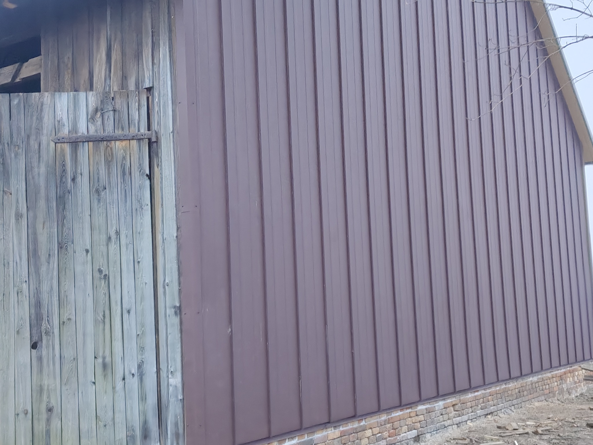 Brązowe, blaszane poszycie dachu budynku gospodarczego, kontrastujące z drewnianymi, szarymi drzwiami i ceglaną podmurówką. Widoczne detale mocowania blachy.