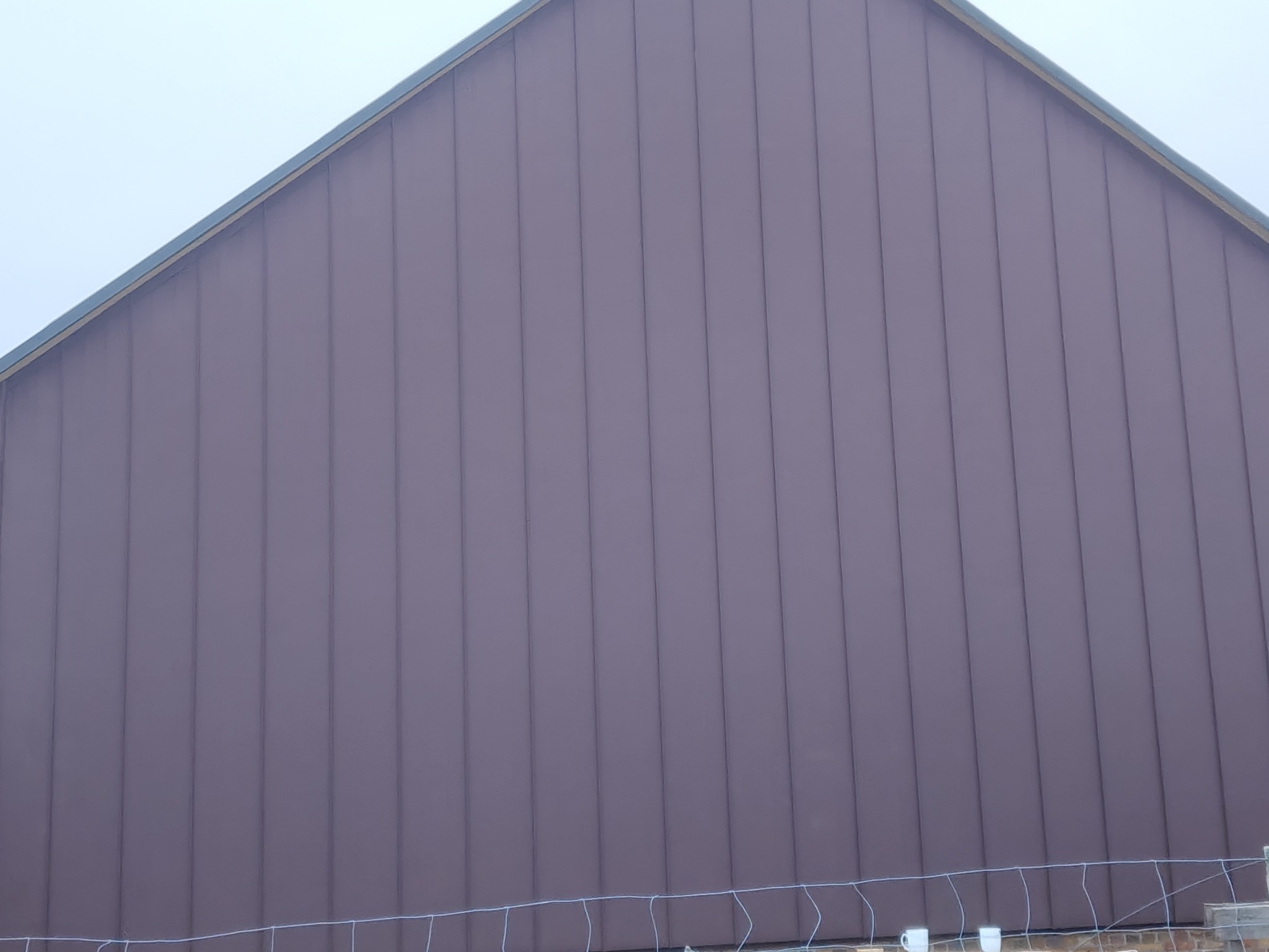 Elewacja szczytowa budynku pokryta panelami w odcieniach brązu, widoczne pionowe łączenia paneli, fragment ogrodzenia w dolnej części kadru.