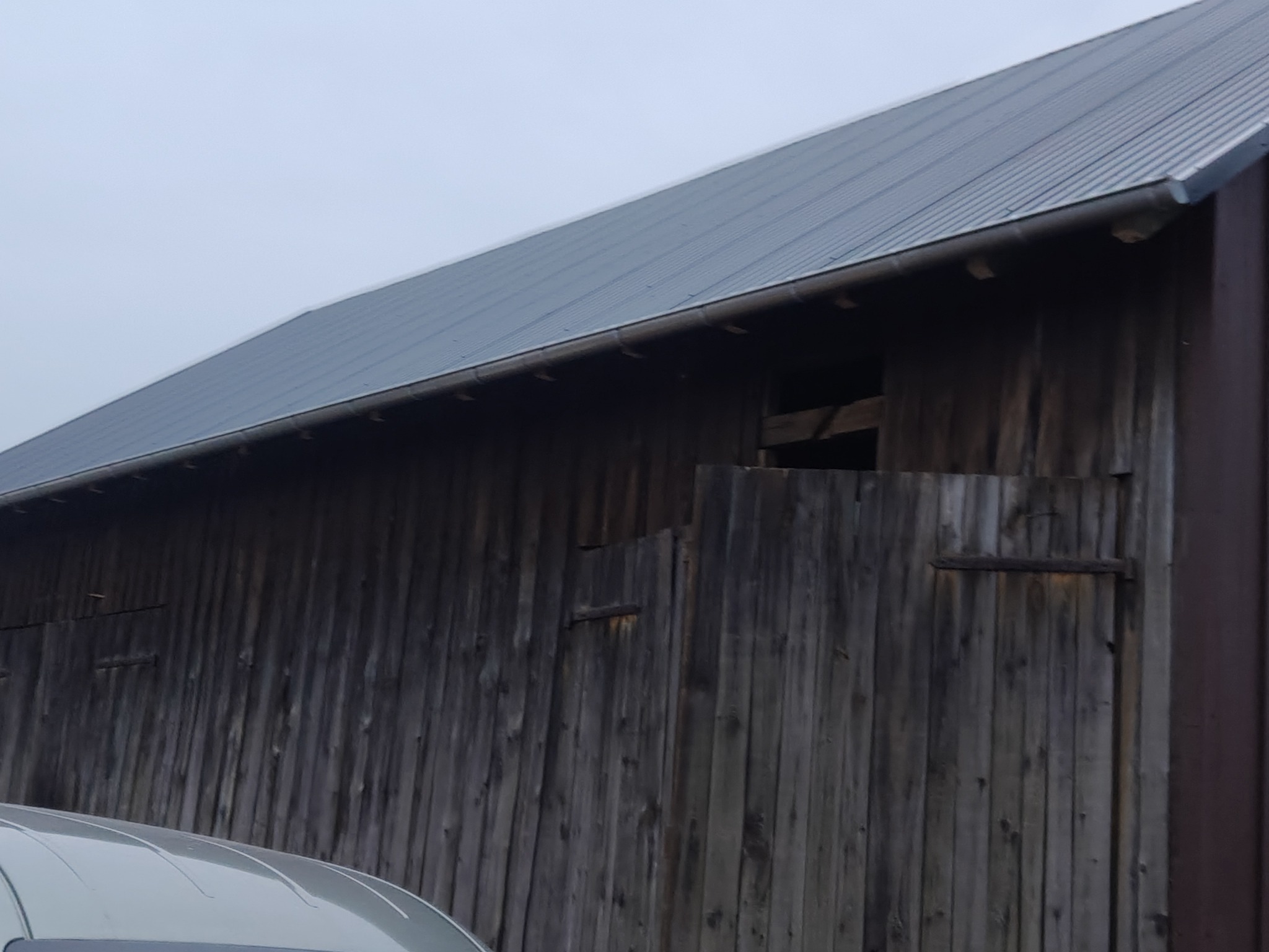 Szary dach z blachy trapezowej na drewnianym budynku gospodarczym z zamontowanymi rynnami. Widoczne deski elewacyjne i fragment pojazdu w dolnej części kadru.