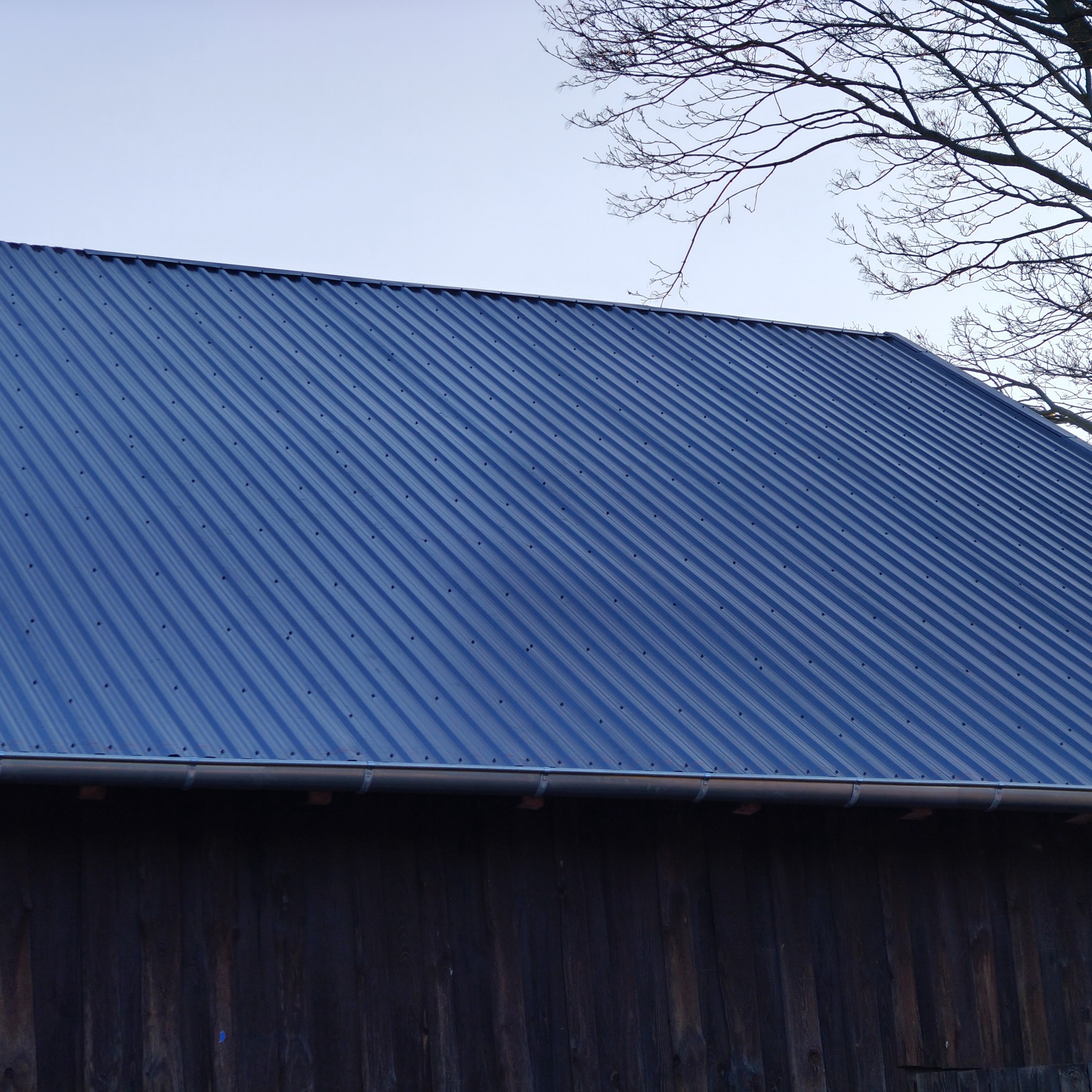Niebieski dach z blachy trapezowej z zamontowaną rynną, widoczny fragment drewnianej ściany budynku i gałęzie drzewa na tle jasnego nieba.