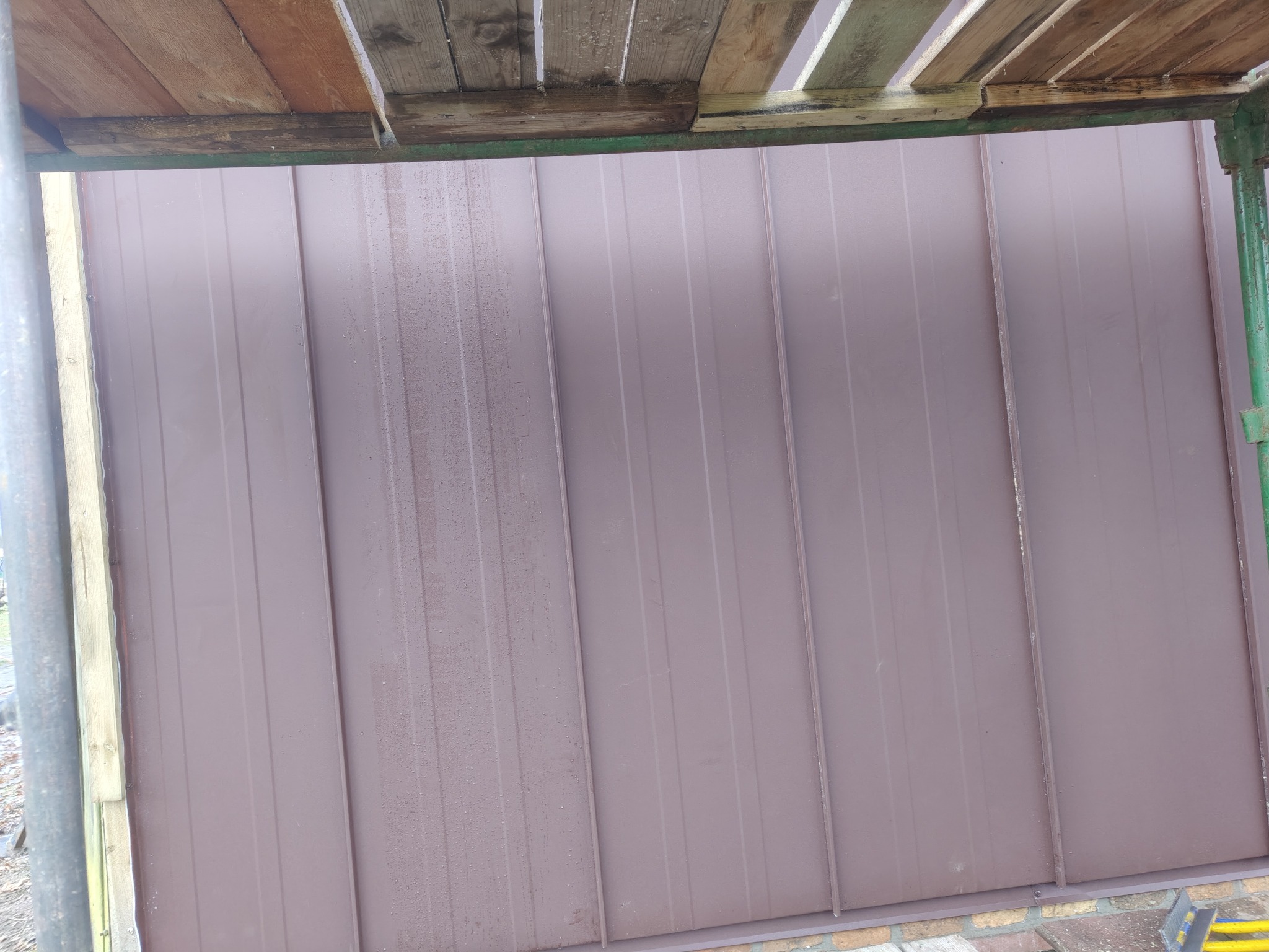 Nowe, brązowe panele blaszane na dachu, widoczne ze rusztowania. Świeżo położone, z delikatnymi śladami wilgoci. Ujęcie z dołu, podkreślające regularność i estetykę.