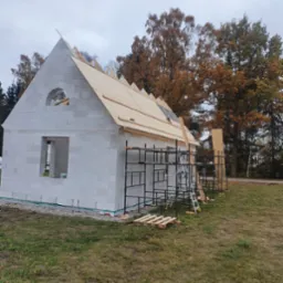 Wznoszenie ścian parterowego budynku z bloczków betonu komórkowego, widoczne rusztowania i elementy konstrukcji dachu na tle jesiennego krajobrazu.