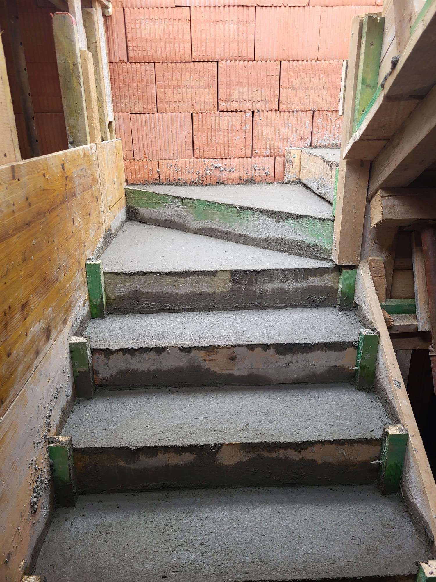 Surowe betonowe schody w szalunku z drewnianych desek i zielonych podpór, ceglana ściana w tle, etap budowy.