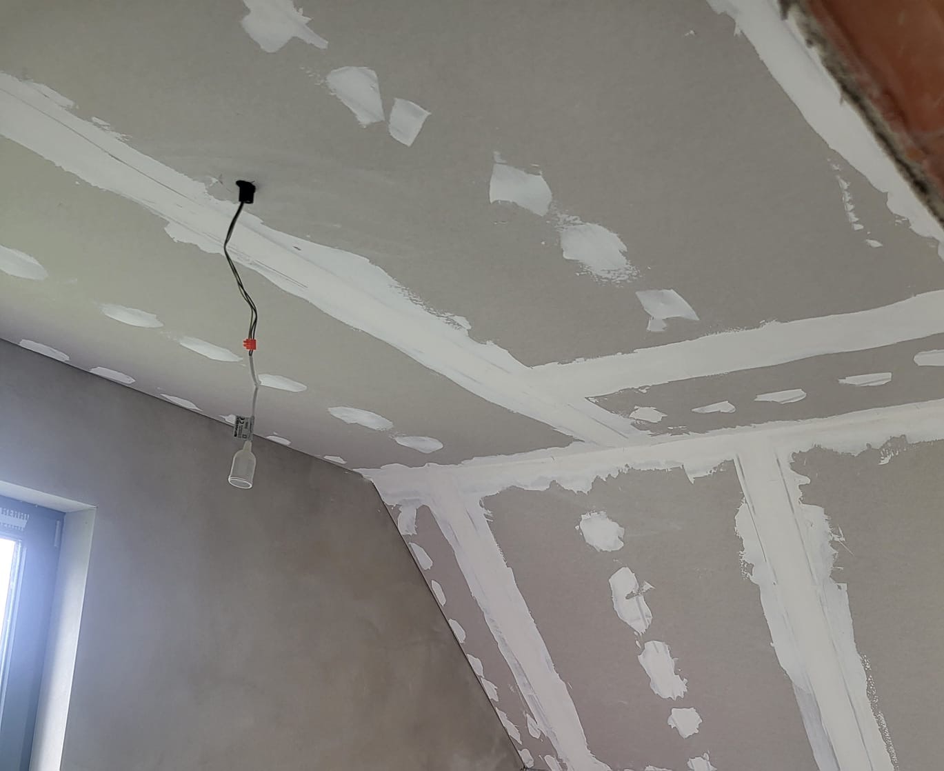 Wnętrze w trakcie prac wykończeniowych: szpachlowana ściana i sufit pod skosem z widocznymi łączeniami płyt gipsowo-kartonowych, prowizoryczne oświetlenie z żarówką na kablu.