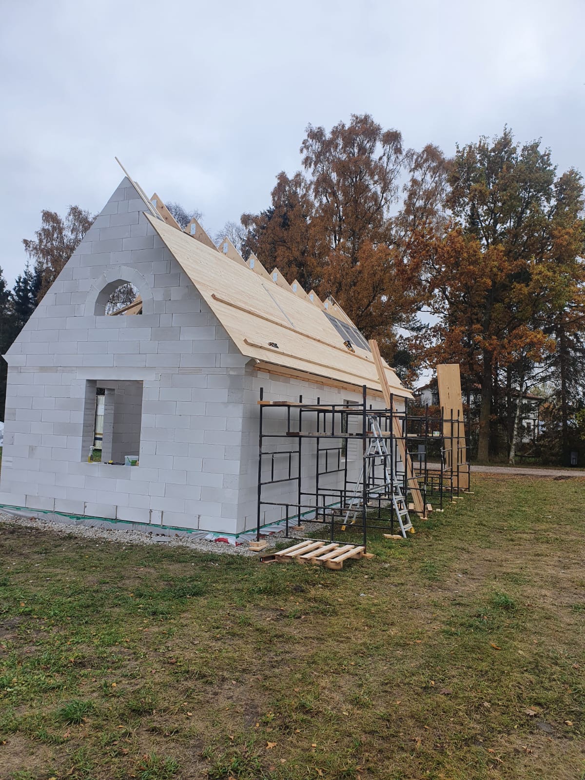 Wznoszenie ścian parterowego budynku z bloczków betonu komórkowego, widoczne rusztowania i elementy konstrukcji dachu na tle jesiennego krajobrazu.