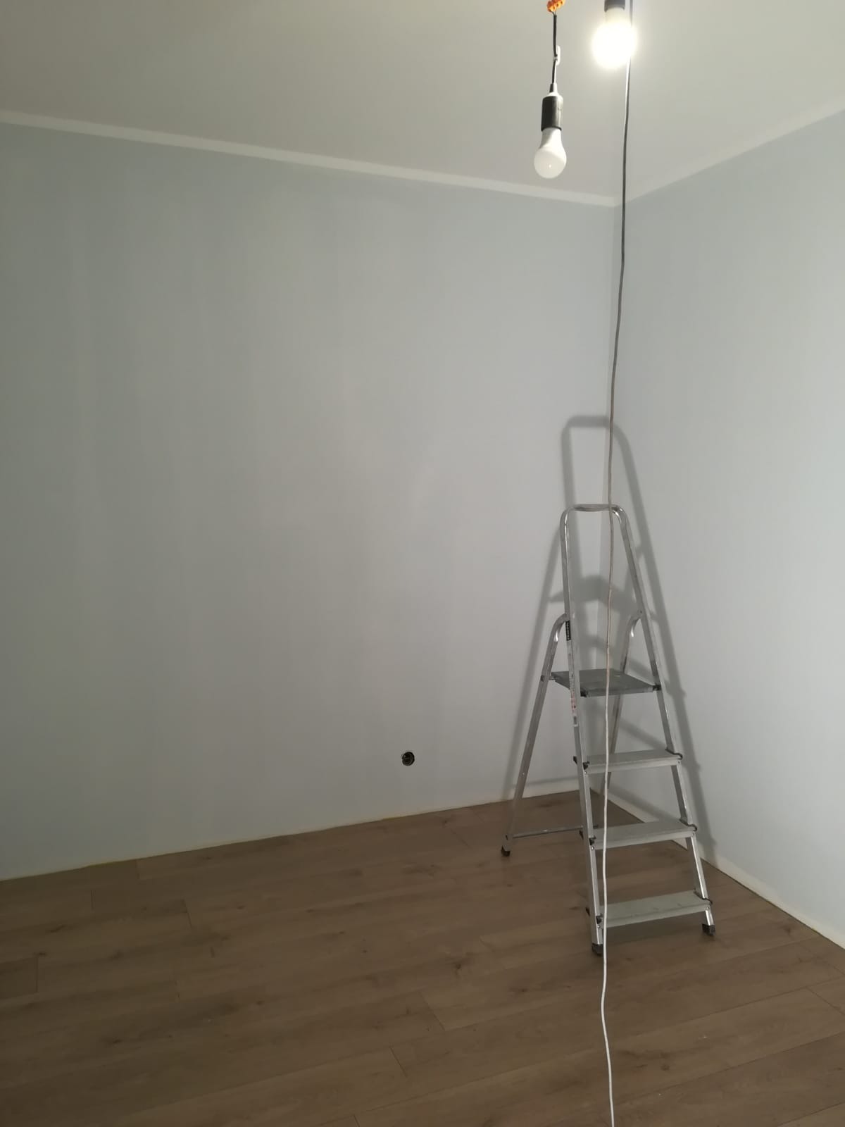 Pokój po pomalowaniu ścian na jasny szaro-niebieski kolor, z widoczną drabinką malarską, drewnianą podłogą i prowizorycznym oświetleniem z żarówką.