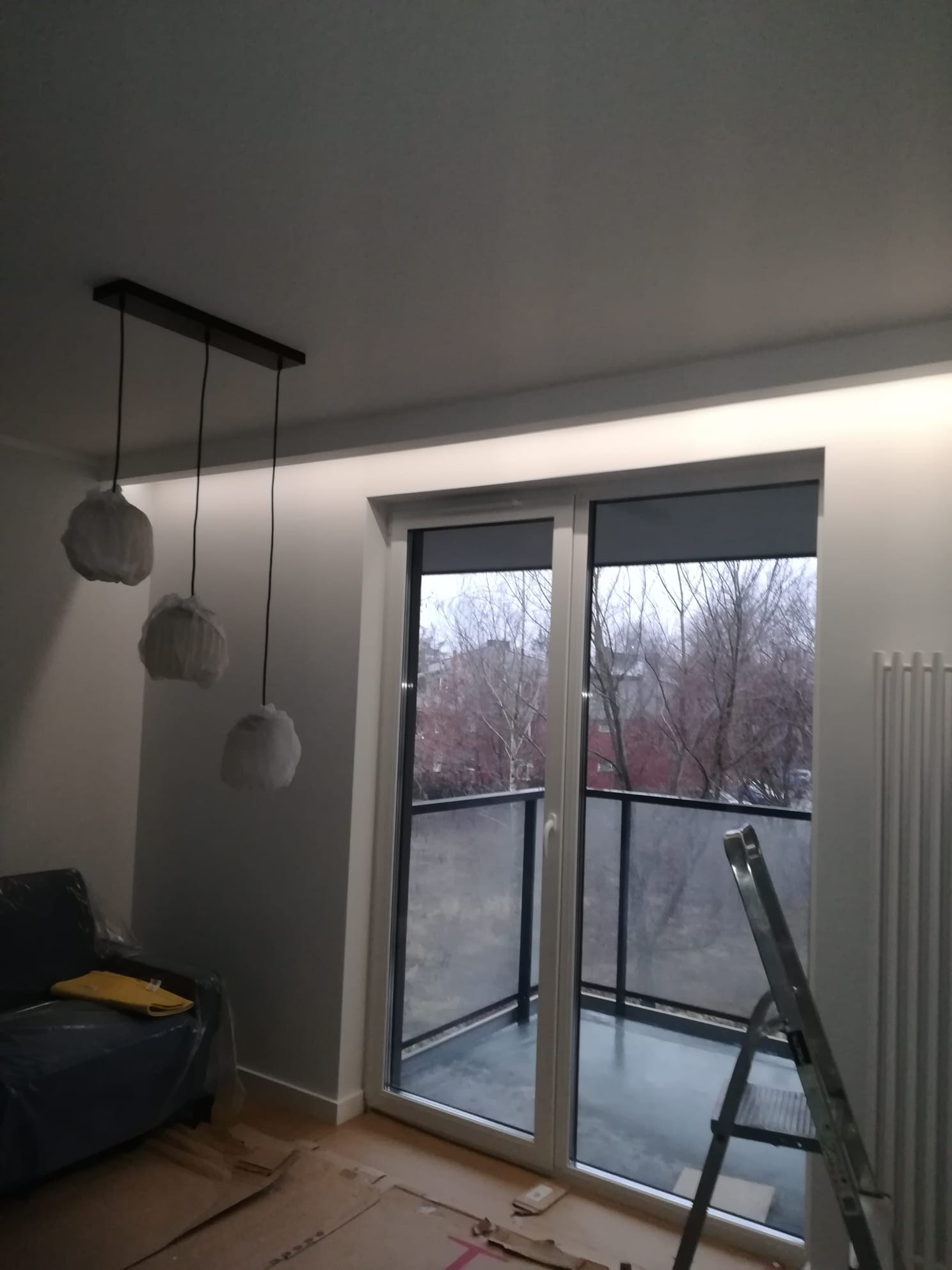 Wnętrze w trakcie wykańczania: widoczne osłonięte lampy wiszące na czarnych kablach, okno balkonowe z szarą roletą, drabina oparta o ścianę i zabezpieczona folią sofa.